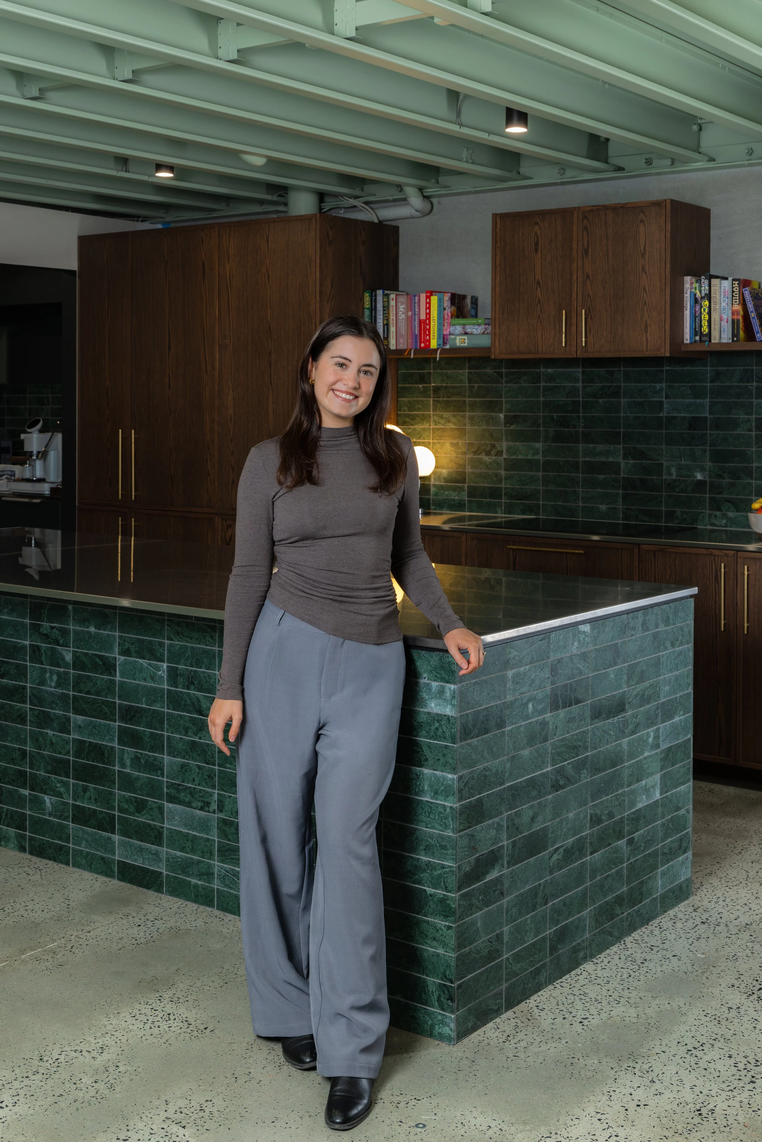 A woman smiling and standing in a modern kitchen with dark green tiled counter, wooden cabinets, and open shelving with books. She is wearing a gray turtleneck and gray wide-leg trousers. The ceiling has green exposed beams with lighting.