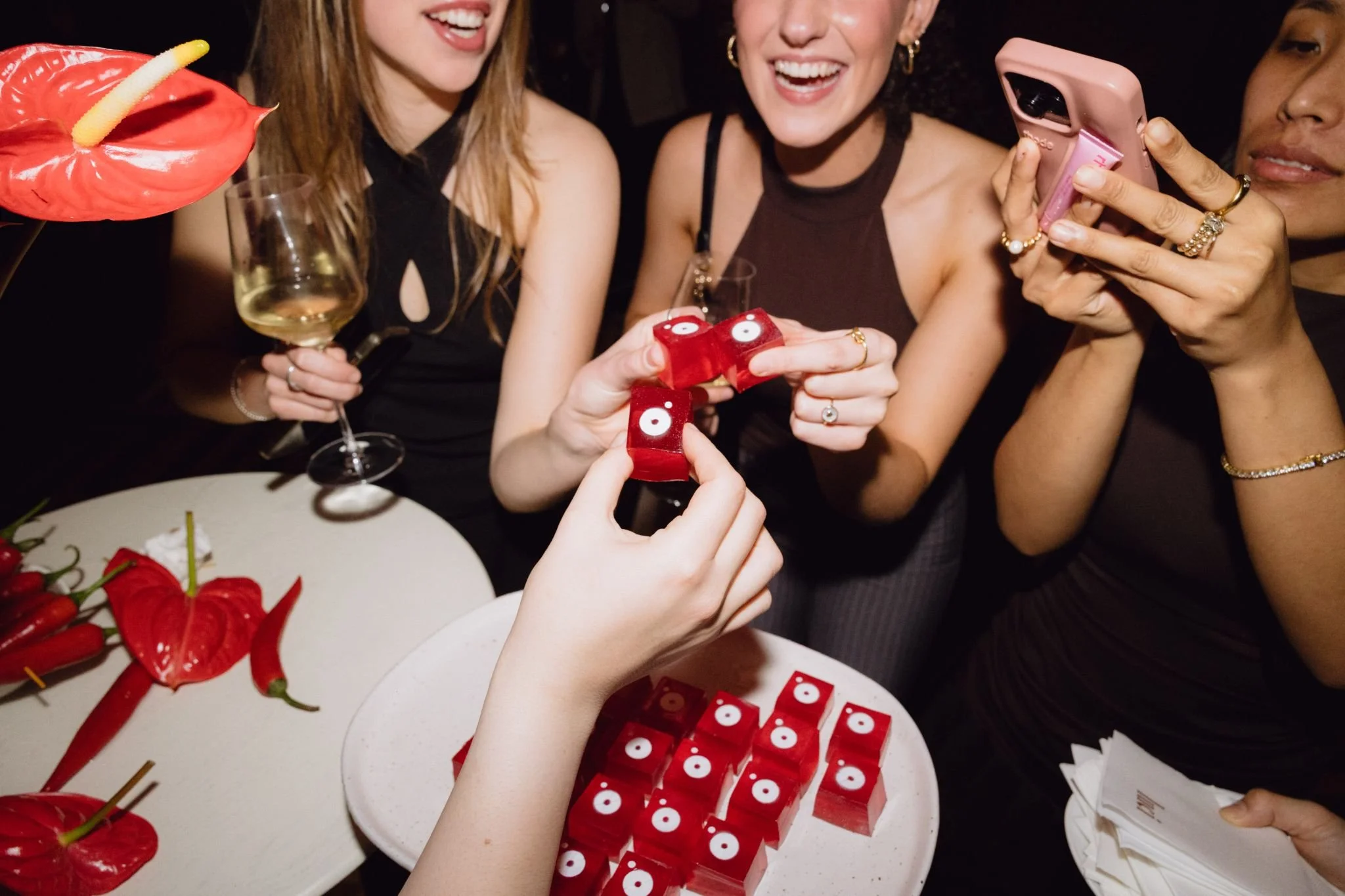 A group of women celebrating at a party, smiling and enjoying themselves. They are holding glasses of white wine, and there is a plate of red flower-shaped desserts with white center decorations on the table.