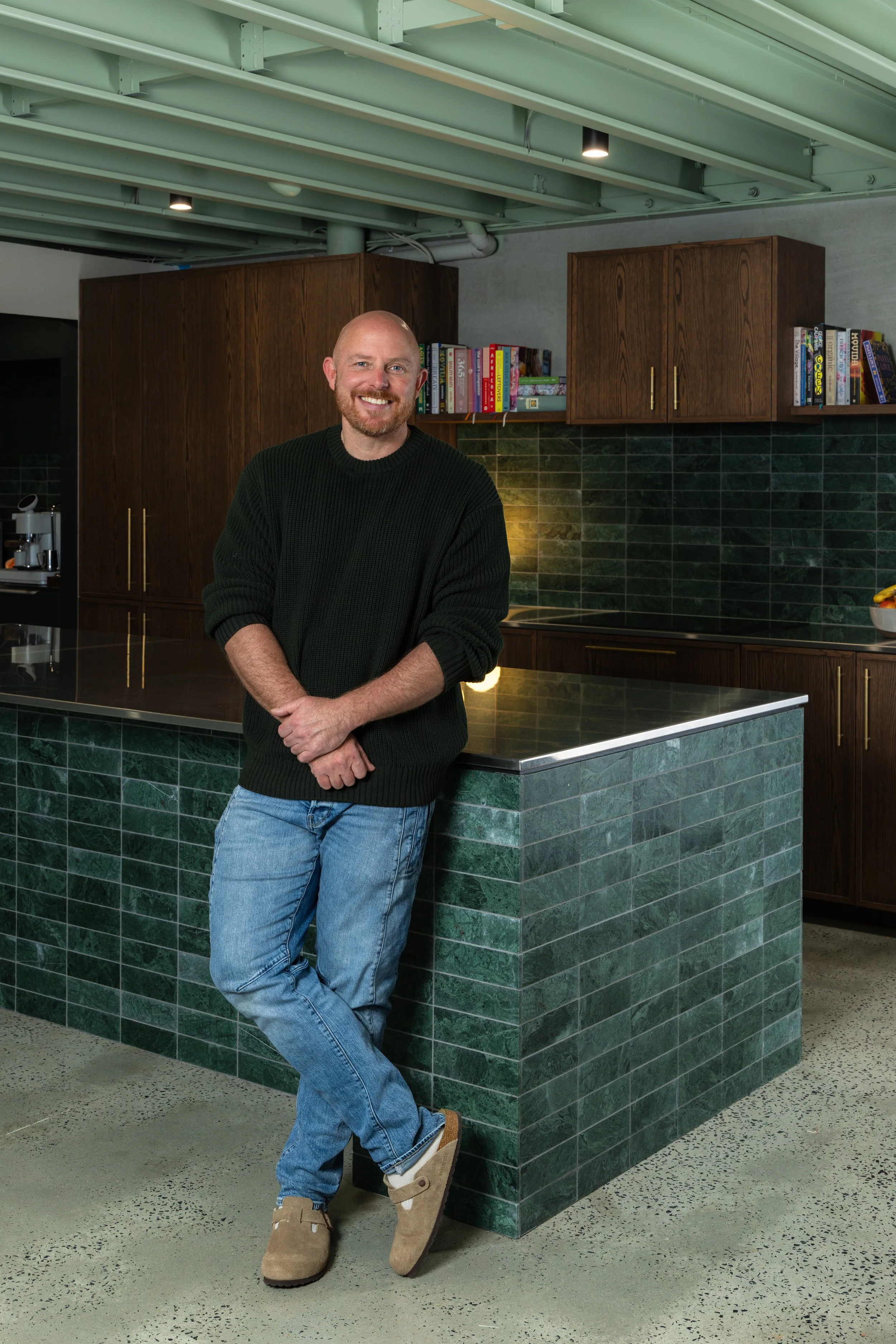 A smiling man with a beard, standing casually at a green tiled kitchen counter with cabinets and books in the background.