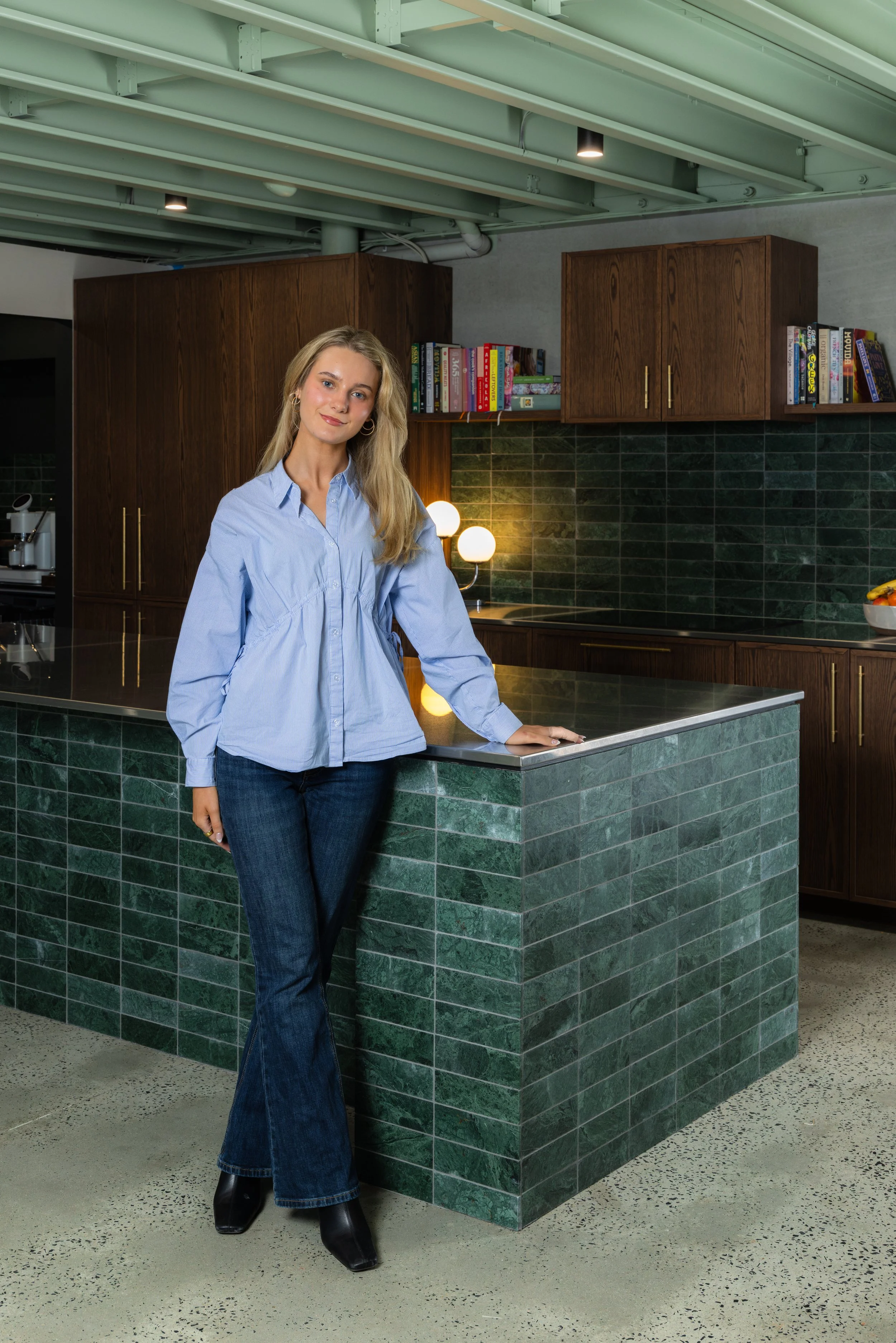 A young woman with long blond hair, wearing a light blue button-up shirt and dark blue flared jeans, is standing next to a green marble kitchen island in a modern kitchen with wooden cabinets, books, and a green tiled backsplash.