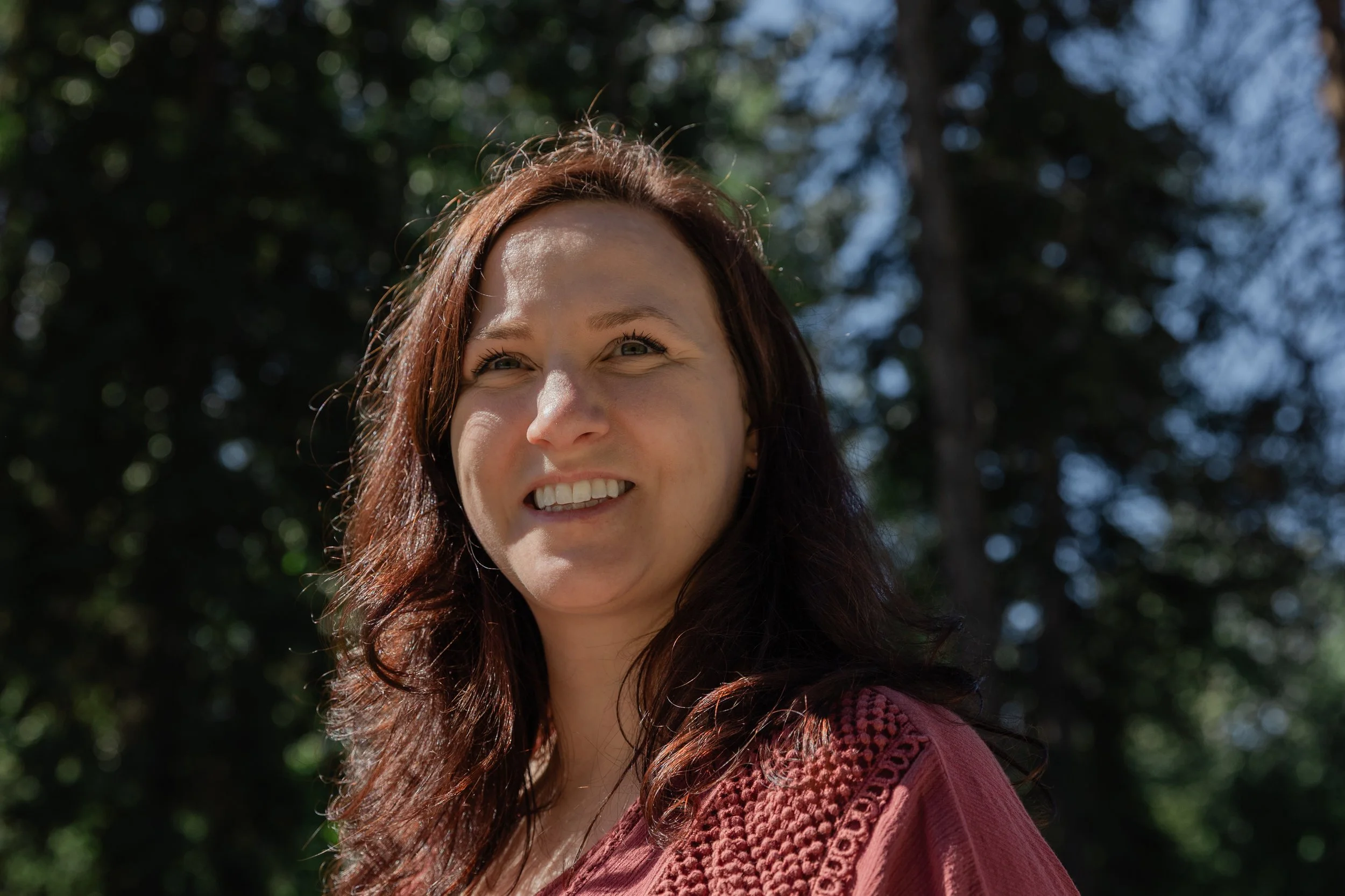 Close-up of a smiling woman with long wavy brown hair, standing outdoors with green trees and blue sky in the background.
