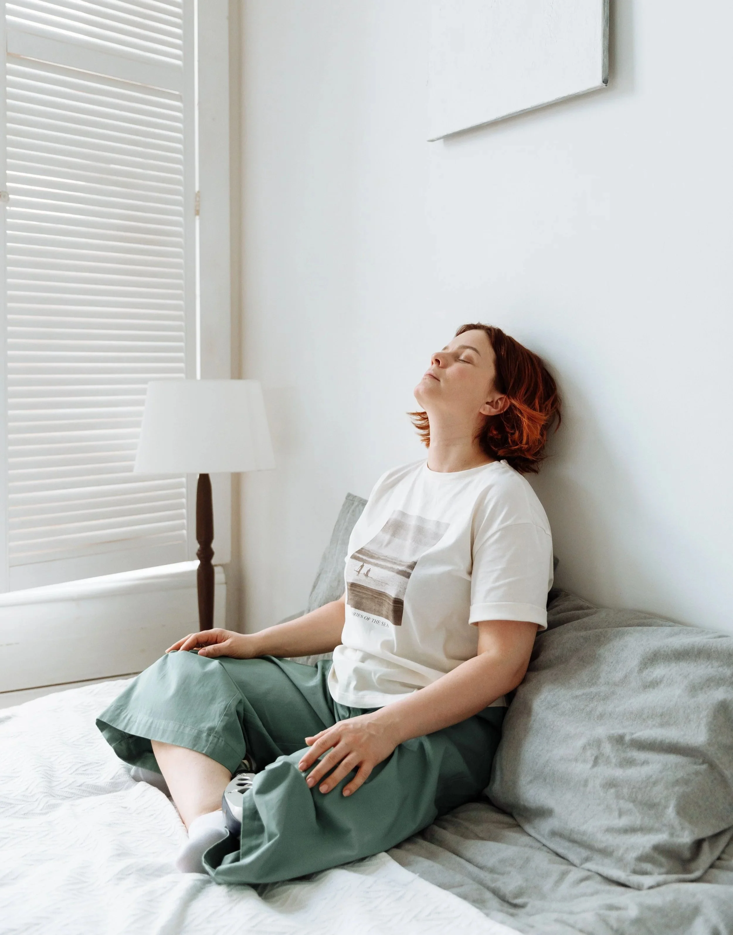 A woman with short red hair relaxing on a bed with gray bedding, sitting upright with her eyes closed, wearing a white t-shirt and green shorts. She is leaning against a gray pillow, next to a nightstand with a white lamp.