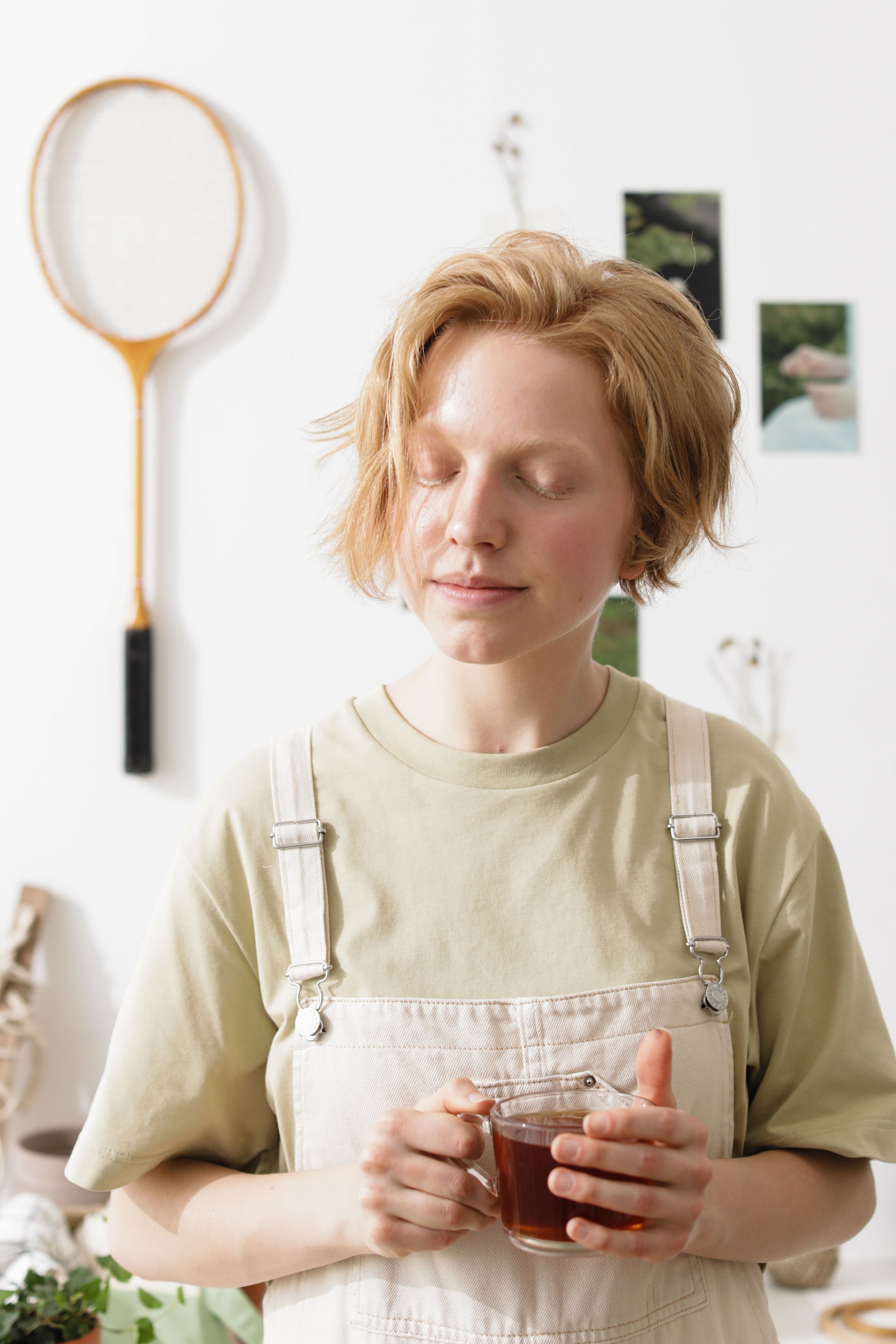 A person with short, wavy blonde hair holding a glass mug of tea or coffee. They are wearing a beige shirt and cream-colored overalls.