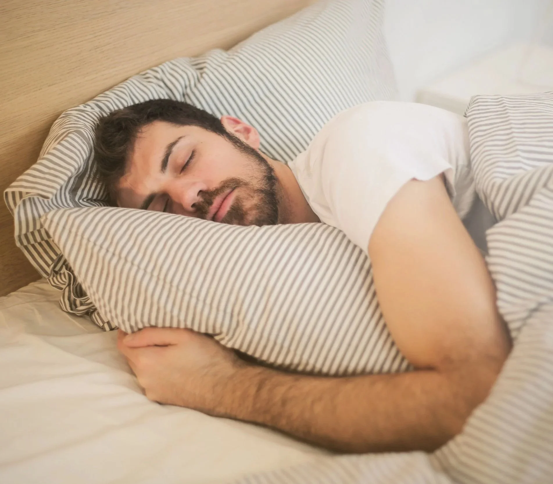 A man sleeping in bed with a striped pillow and matching blanket, resting on a wooden headboard.