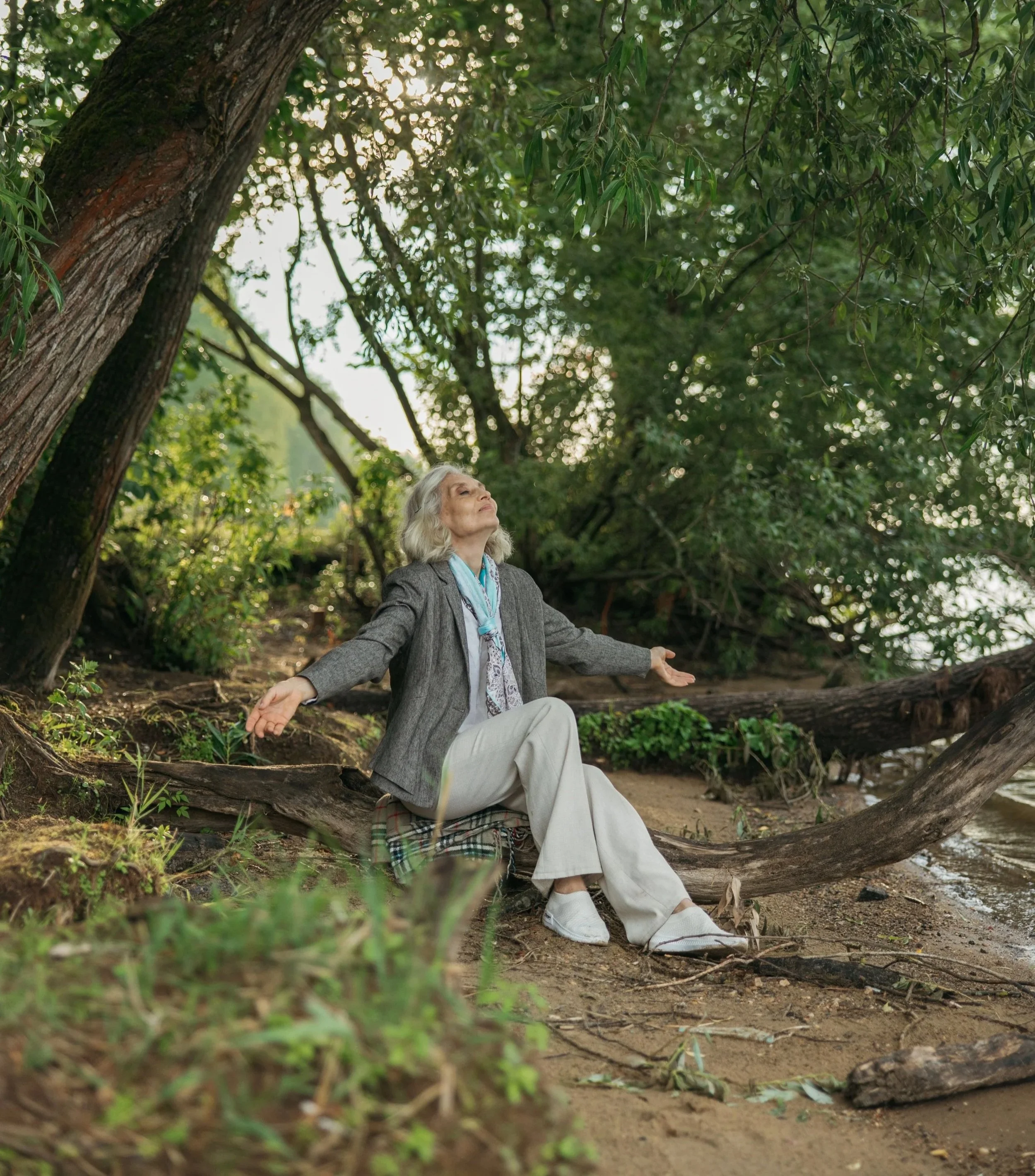 An elderly woman with gray hair sitting outdoors on a fallen log, practicing meditation or mindfulness with her arms open and eyes closed, surrounded by lush green trees and foliage.