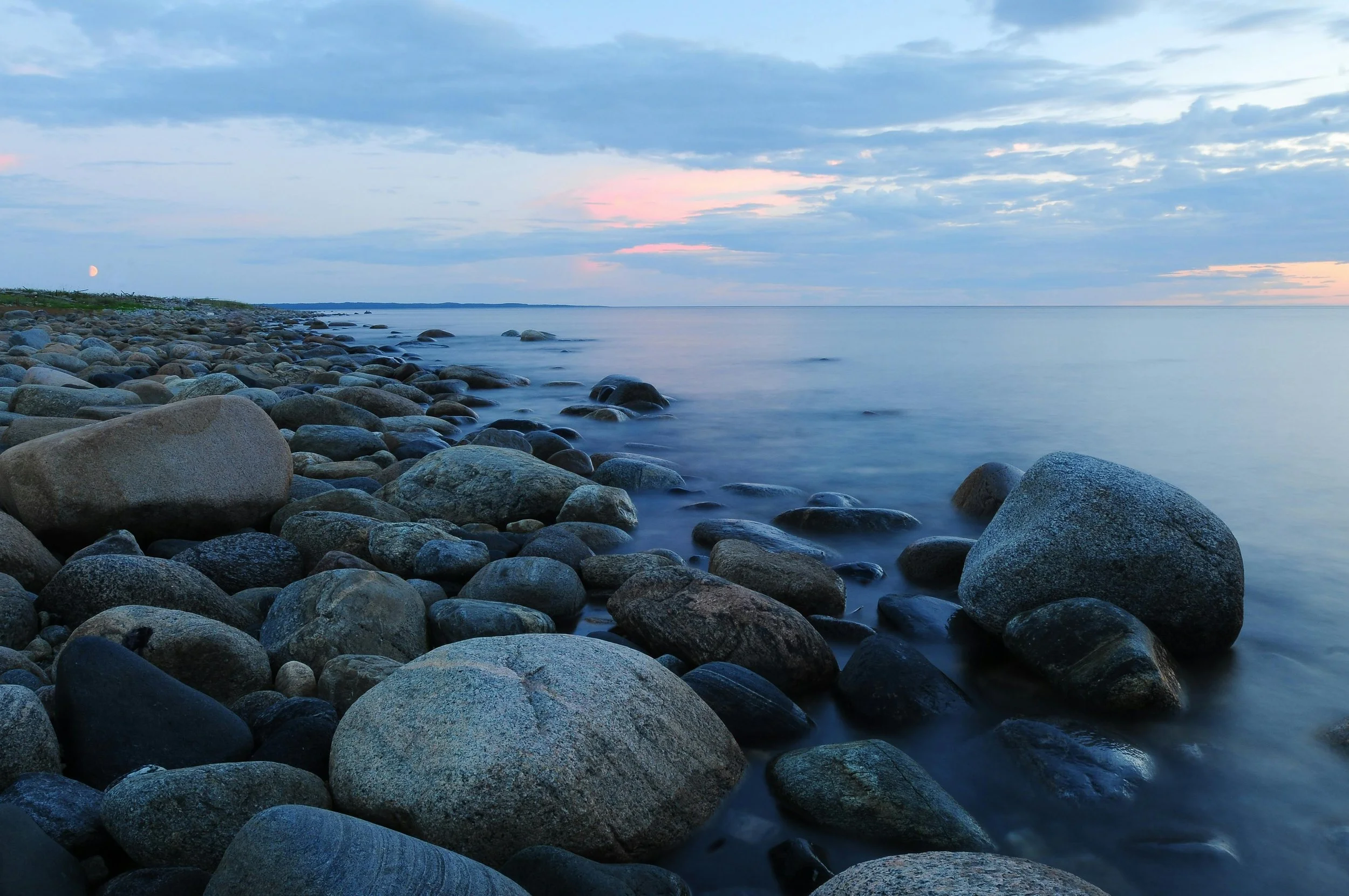A rocky beach with smooth stones along the shoreline, calm water, a distant horizon, and a partly cloudy sky with a visible moon in the early evening or dawn.