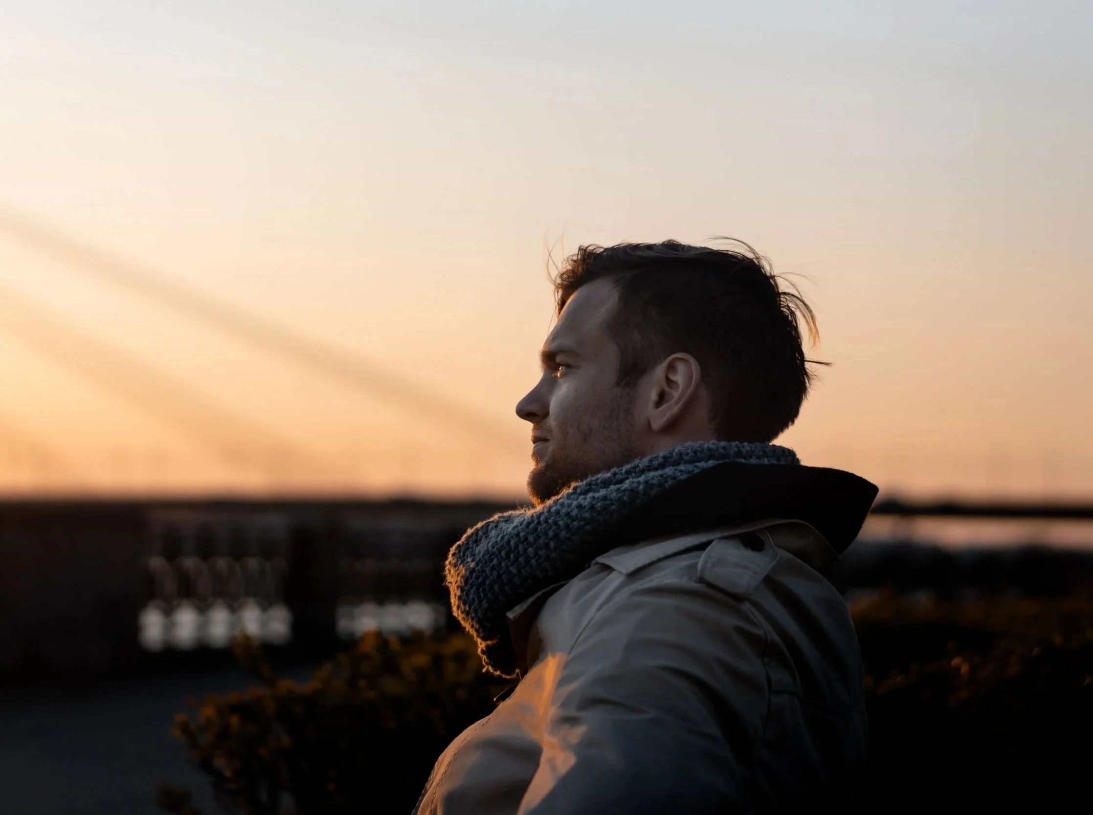 A man with dark hair and a gray scarf looking into the distance during sunset, with a blurred town or cityscape in the background.