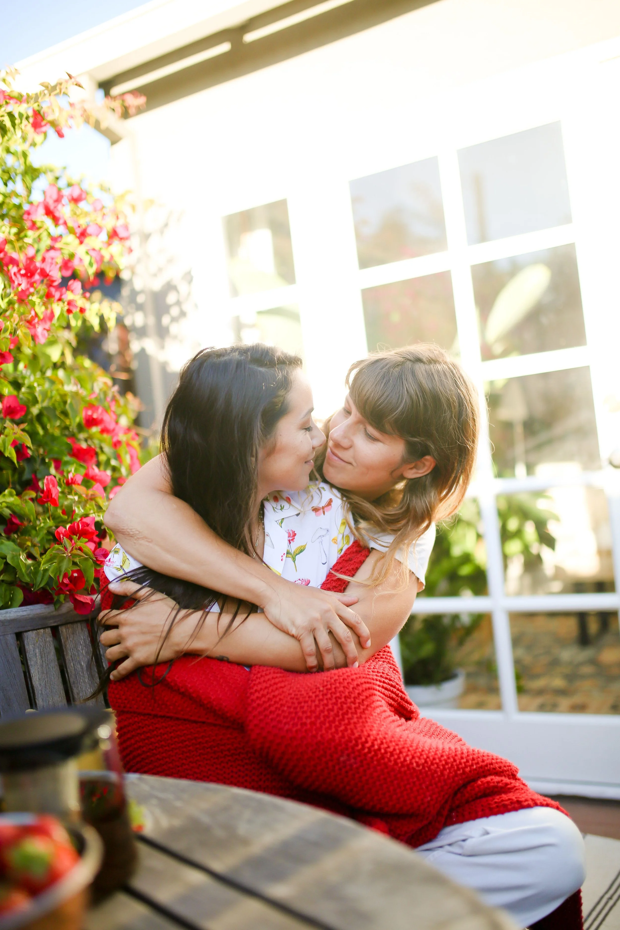 Two young women sit closely together on a bench, embracing and smiling outdoors in a bright setting with a house and blooming plants in the background.