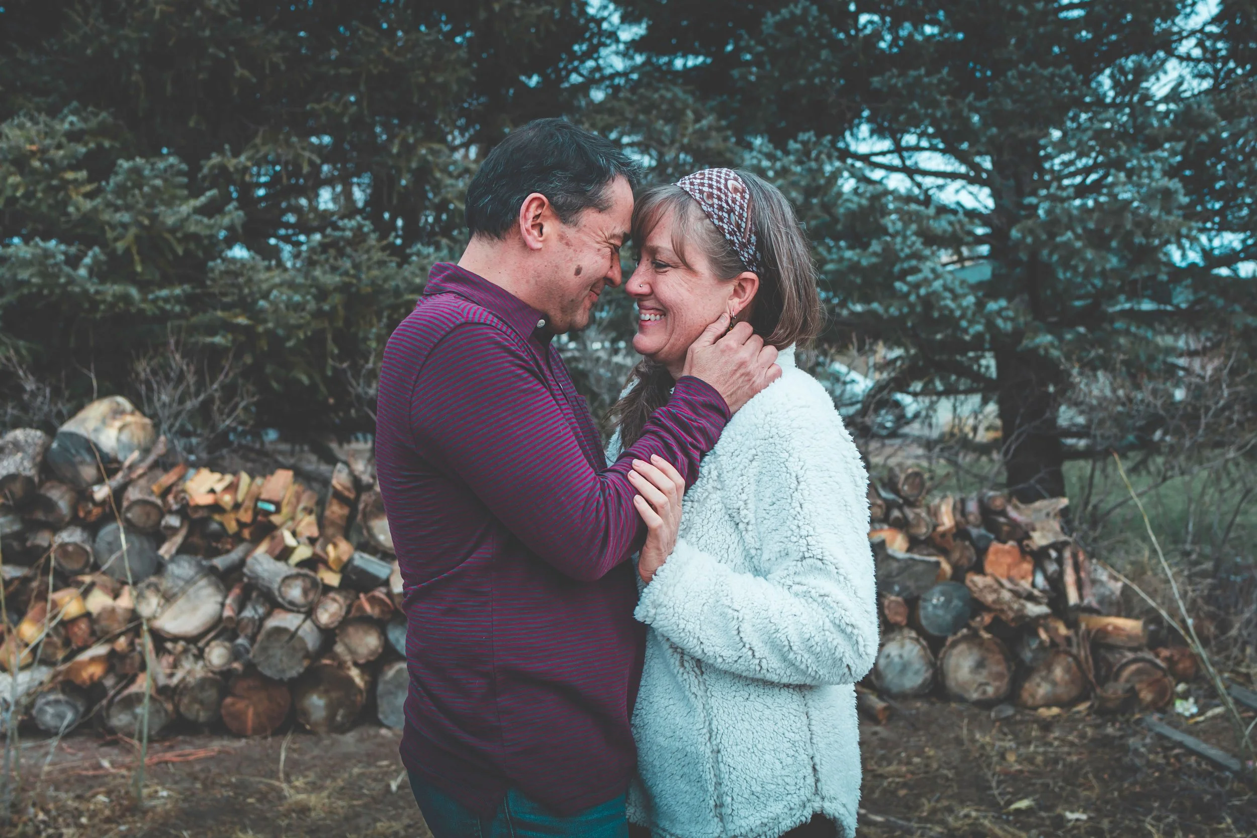 A man and woman are standing close together outdoors, touching foreheads and smiling, with trees and a stacked firewood in the background.