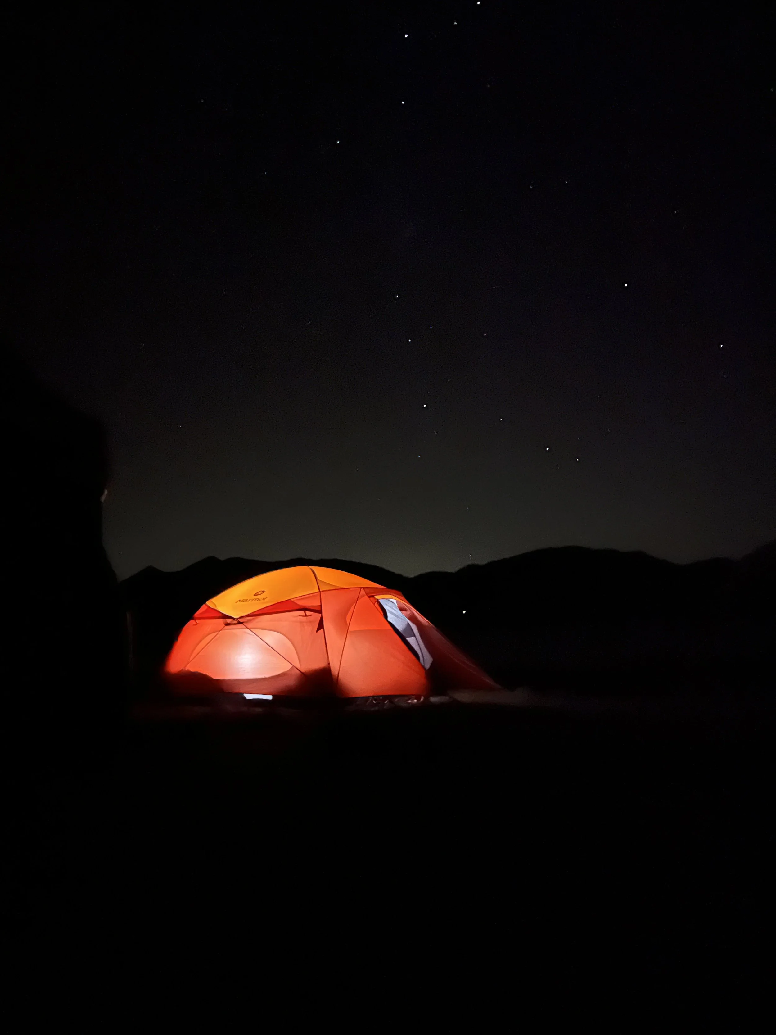 Nighttime scene with a glowing orange tent illuminated from within, set against a dark landscape with mountains and a starry sky.