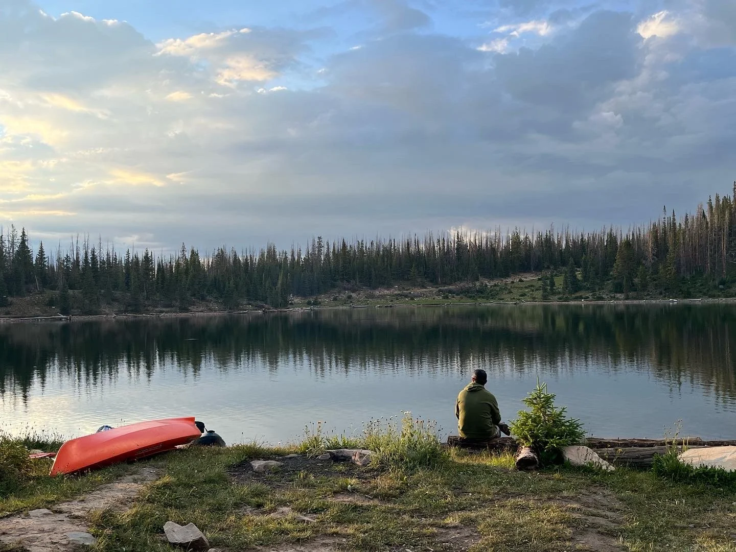 A person sitting on a log by a calm lake with a kayak on the shore, surrounded by a forested landscape and cloudy sky.