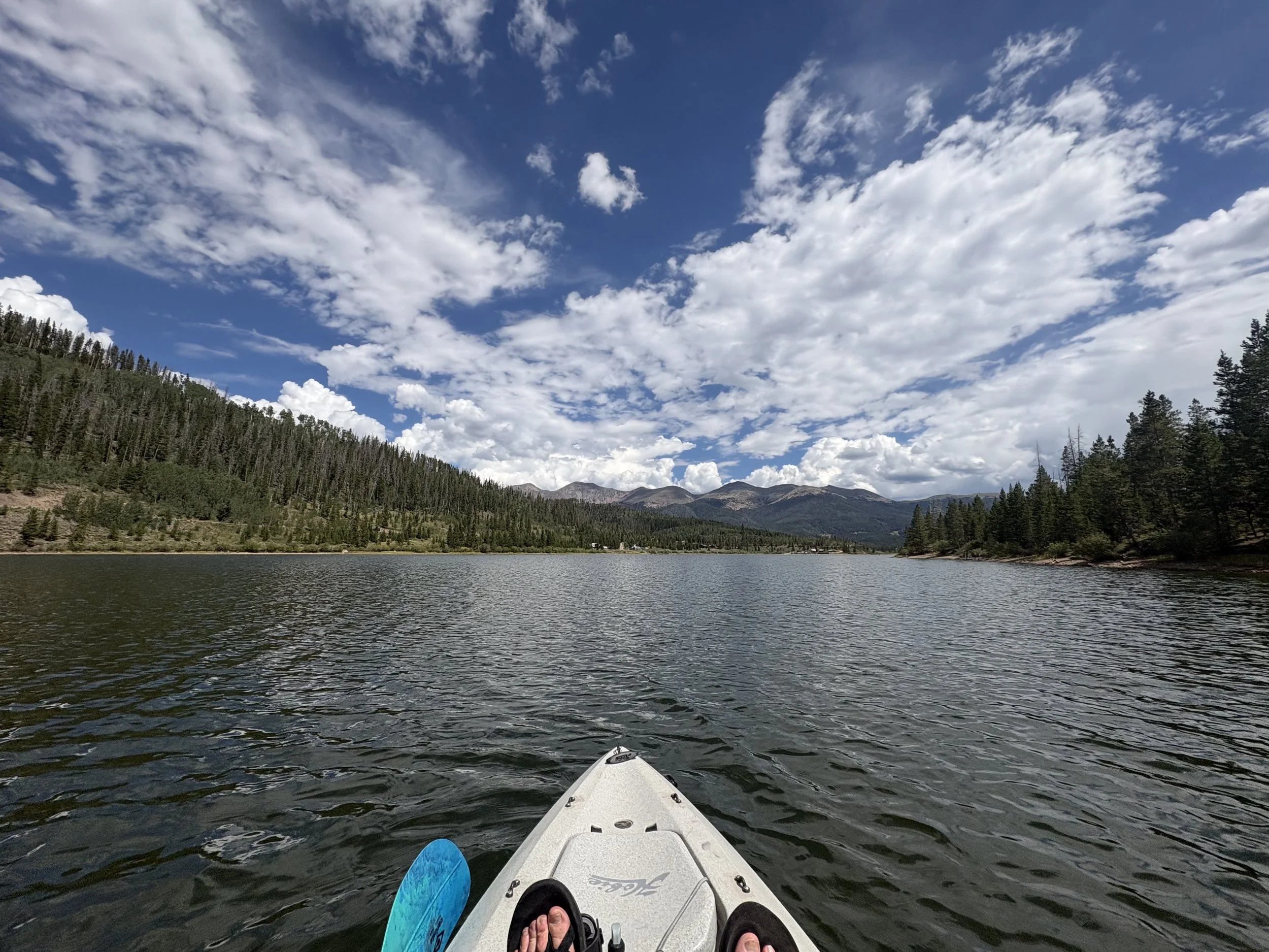 View from a kayak on a lake, with feet wearing sandals, surrounded by forested hills and mountains, under a partly cloudy sky.