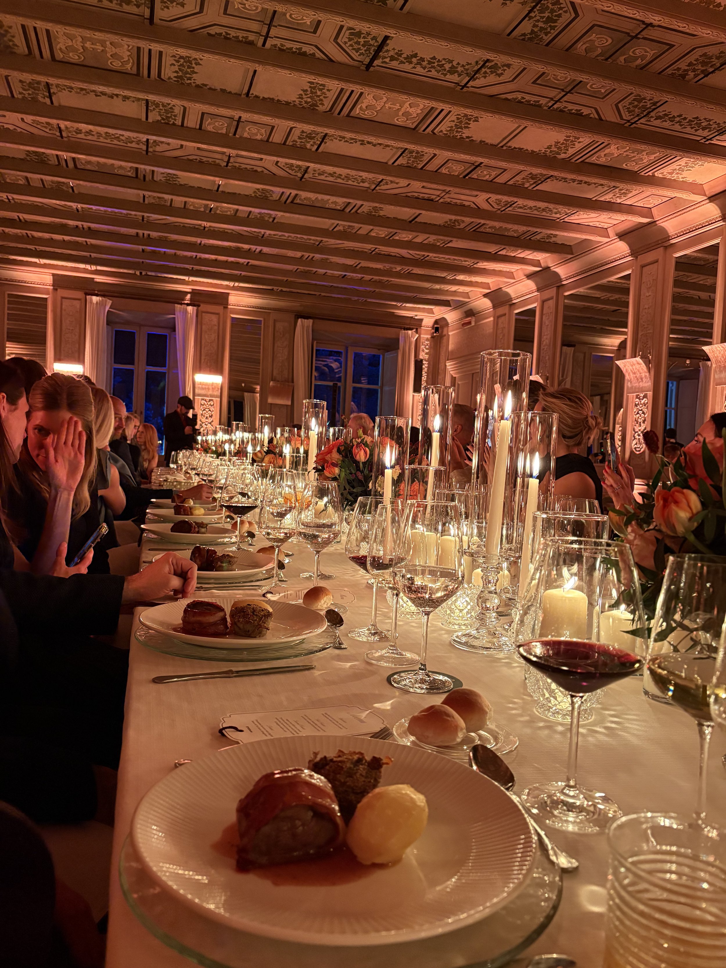 Elegant dinner setting with a long table adorned with lit candles, floral centerpieces, wine glasses, and plated appetizers. Guests are seated along the table in a decorated room with detailed ceiling panels and large windows.