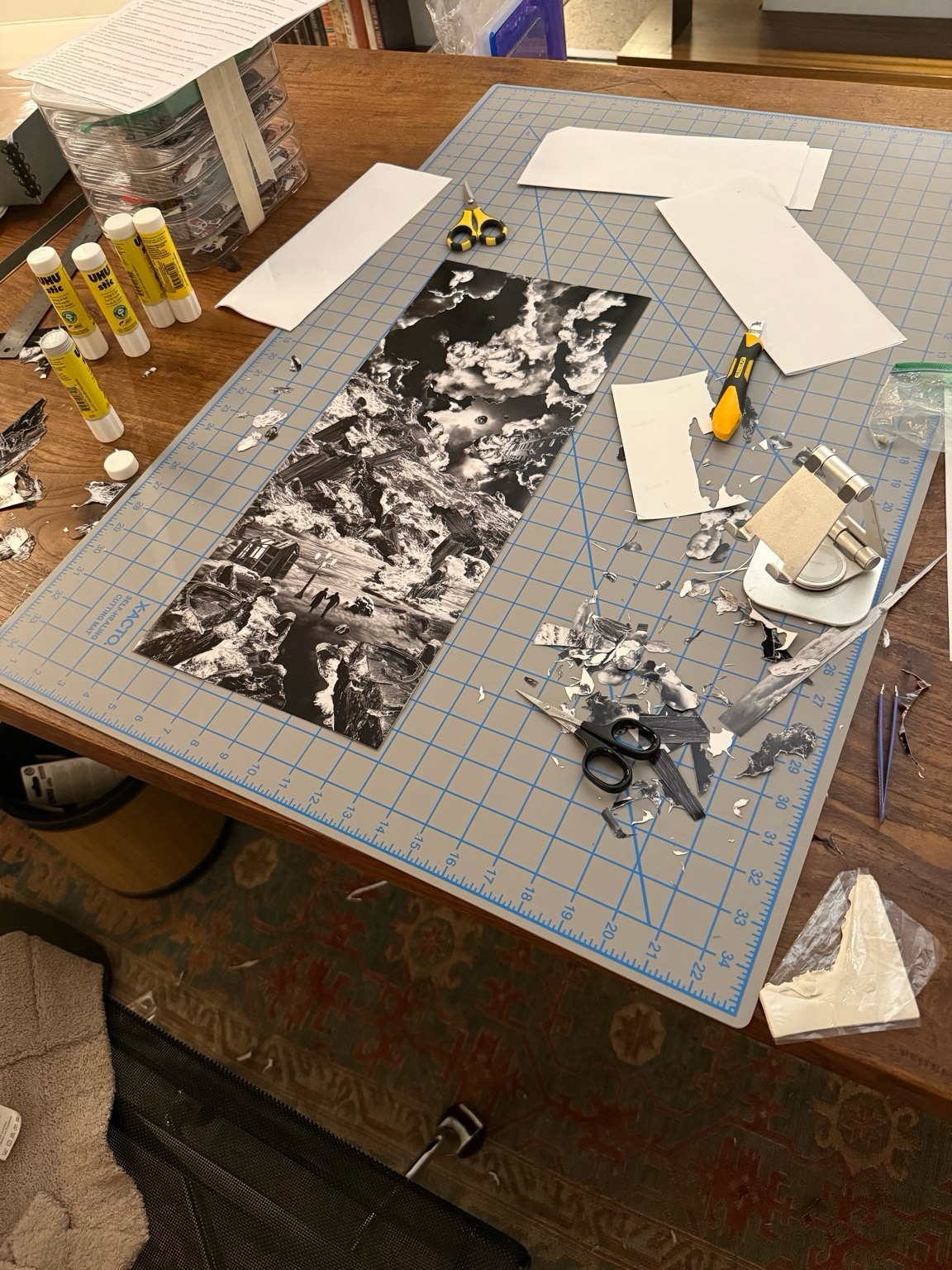 Workstation with art supplies, scissors, glue sticks, a cutting mat, and a black and white landscape collage in process.