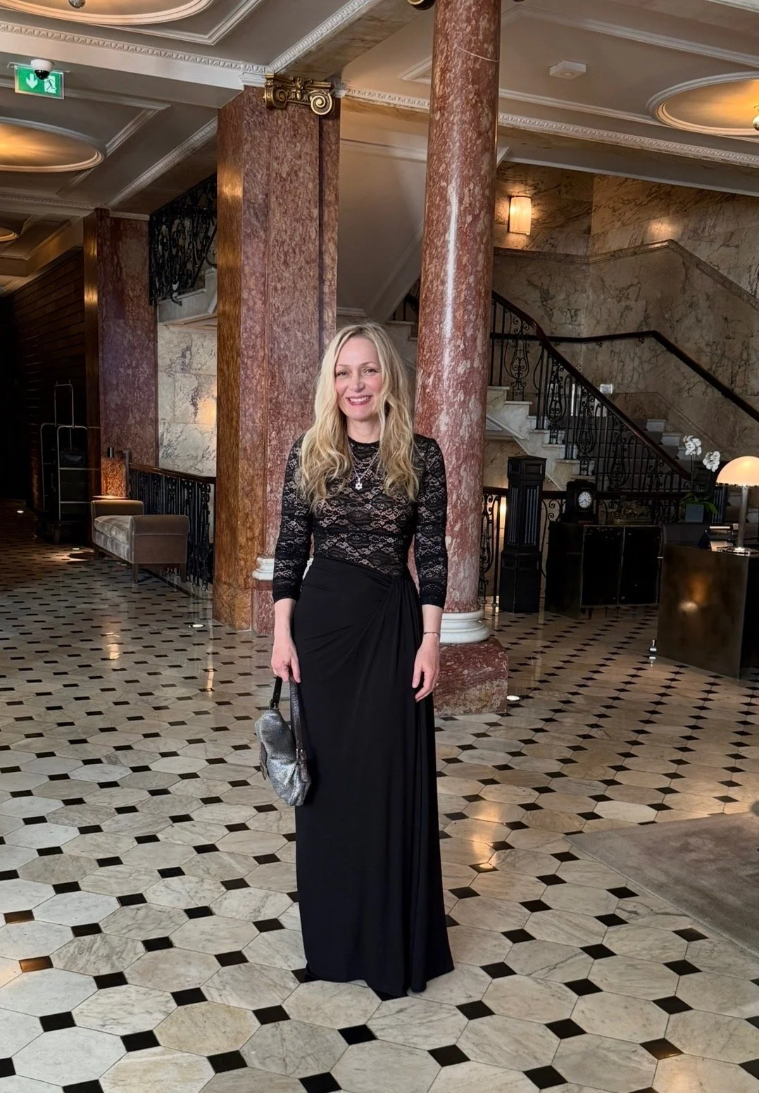 A woman in a black lace top and long black skirt standing in a luxurious hotel lobby with marble pillars, checkered marble floor, and a staircase with an ornate black railing in the background.
