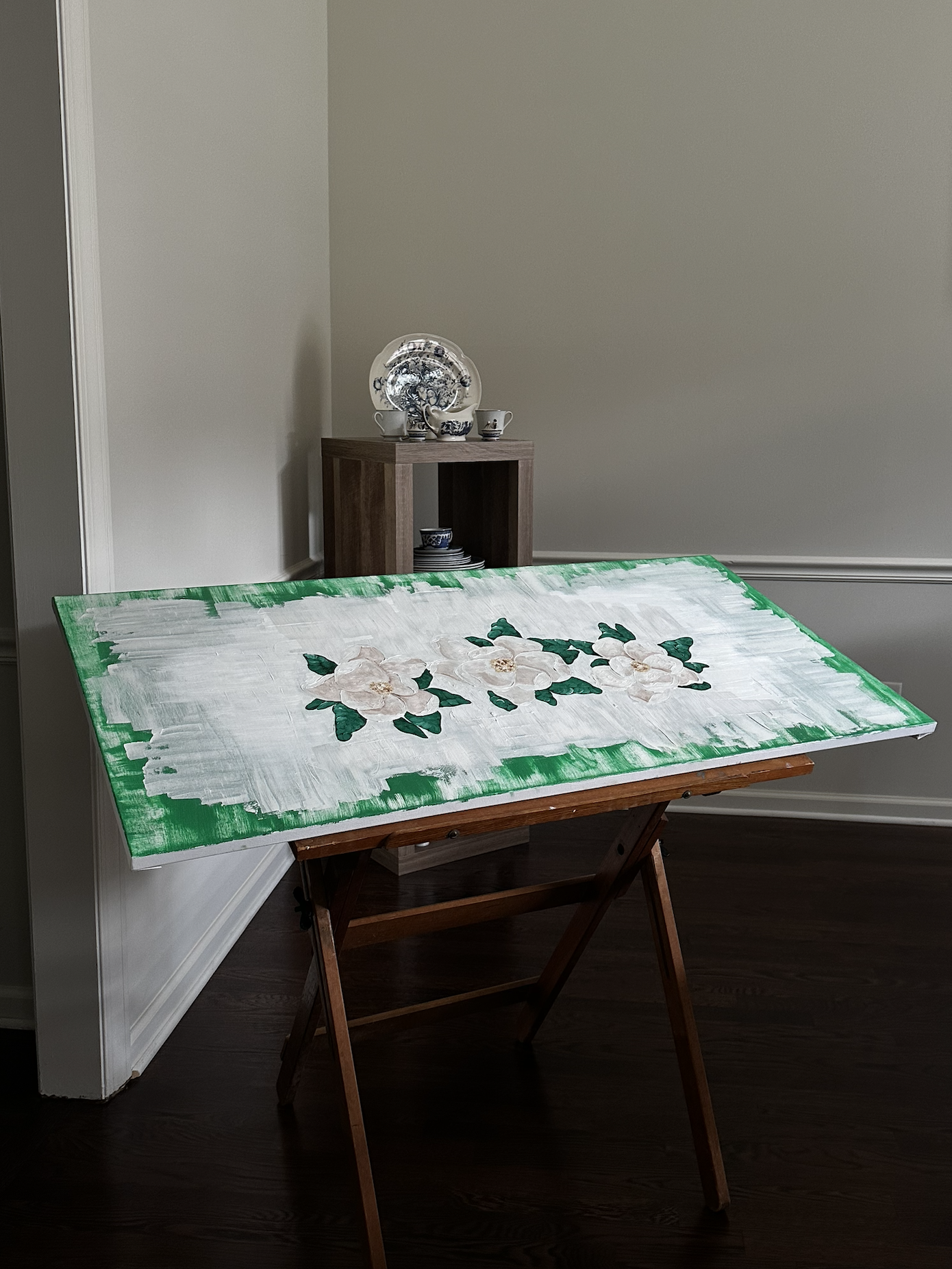 A painting on an easel depicting a floral design with white flowers and green leaves against a white background with green border.