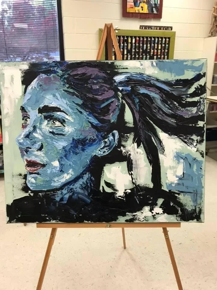 A painting of a woman's face with her hair flowing to the side, featuring bold brushstrokes and a mix of blue, purple, black, white, and red colors, displayed on an easel in an indoor setting.