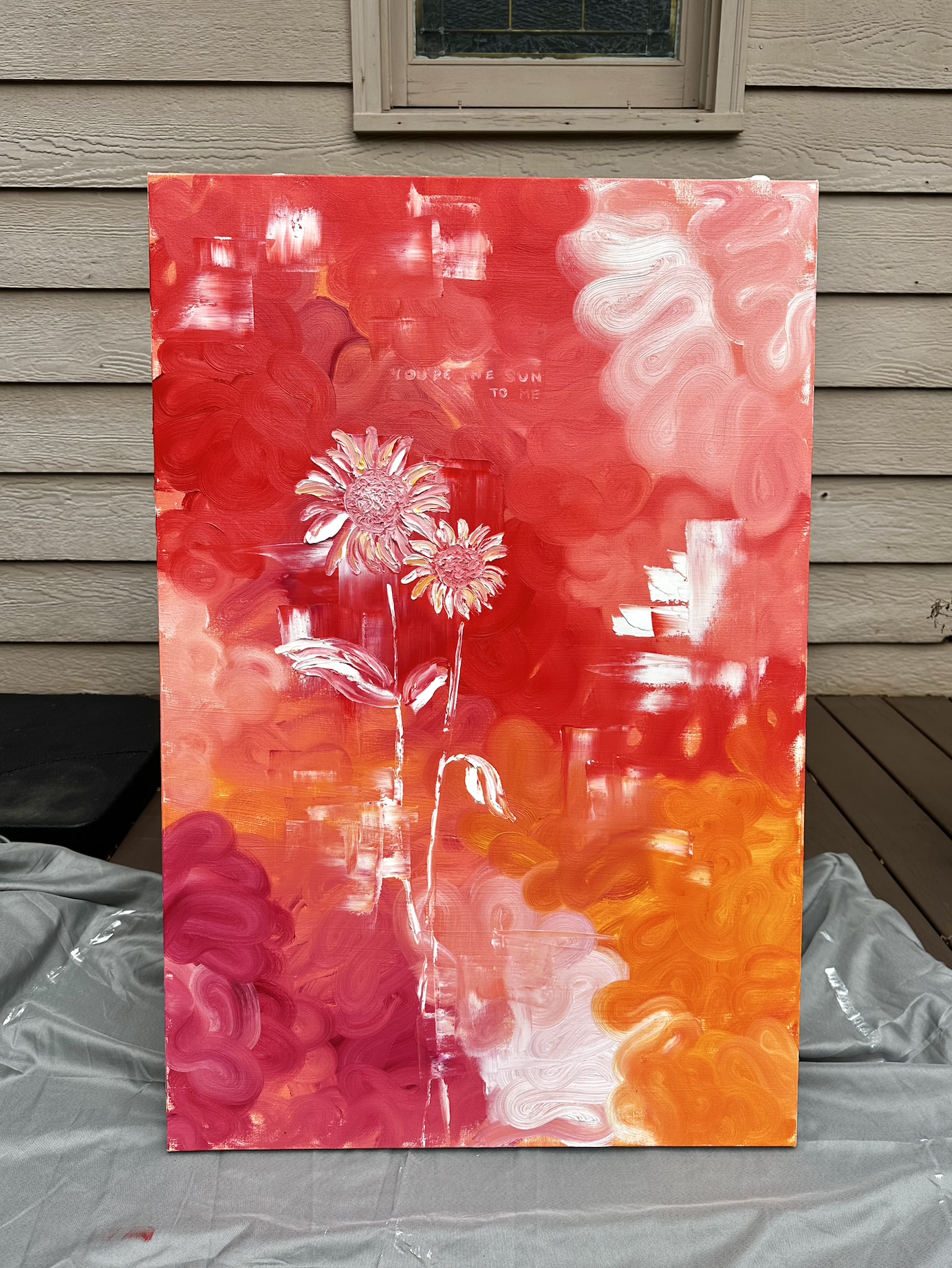 A colorful abstract painting with shades of red, pink, white, and orange, featuring two pink flowers with white and yellow accents in the lower left corner, and text that reads 'You’re the sun to me' in the middle.