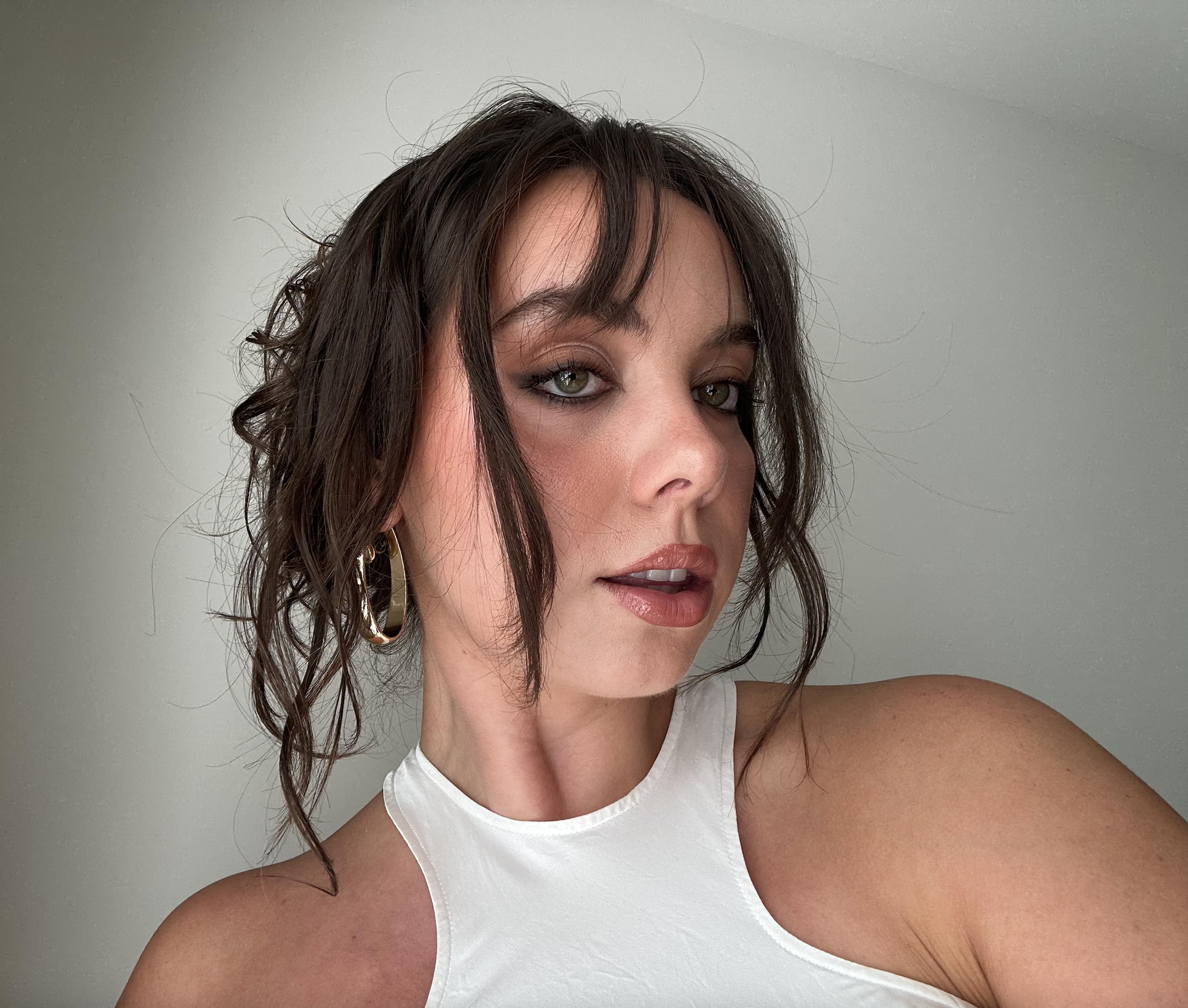 Close-up of a woman with curly brown hair and green eyes, wearing a white sleeveless top, standing against a plain gray background.