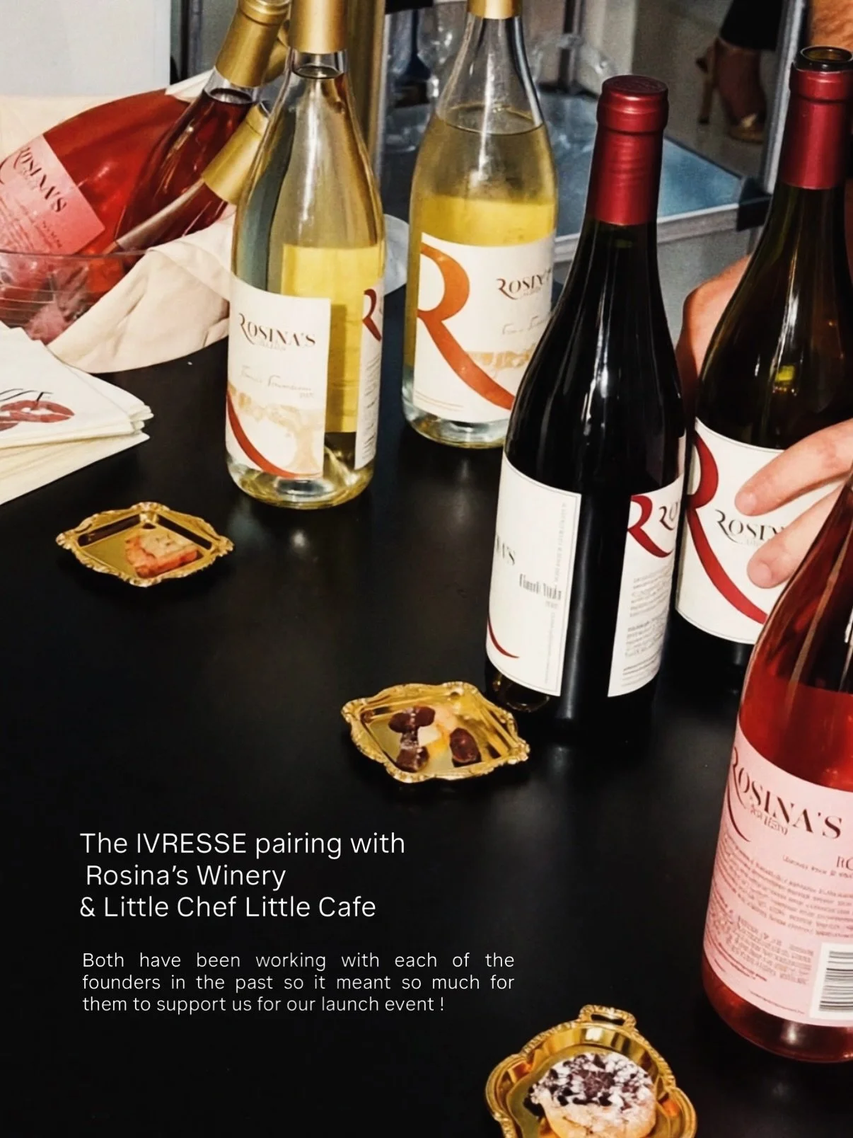 Red and white bottles of wine from Rosina's Winery on a black table, with small gold trays holding chocolates and Hors d’Oeuvres, and a hand reaching for a wine bottle.