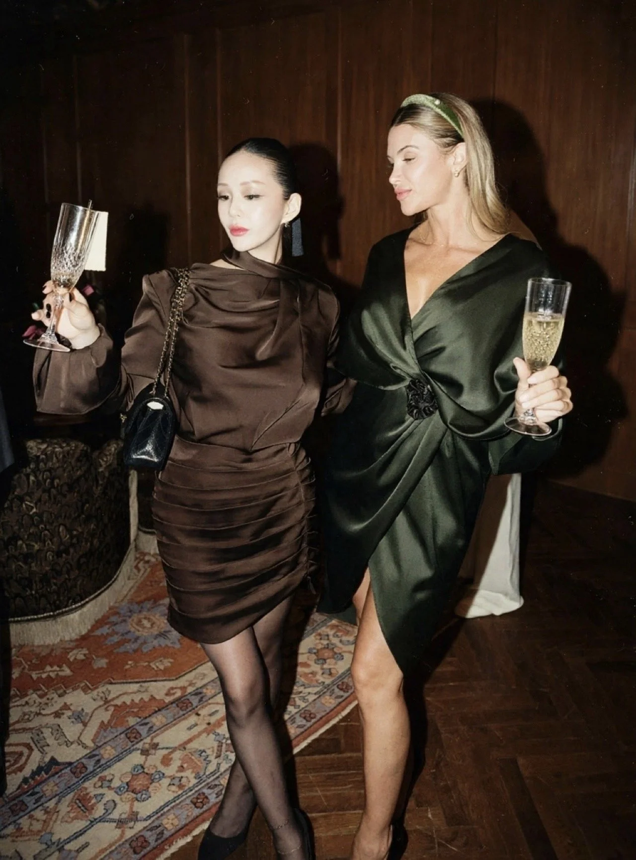 Two women dressed elegantly at a party, holding champagne glasses in a dimly lit room with dark wood walls and a patterned rug.