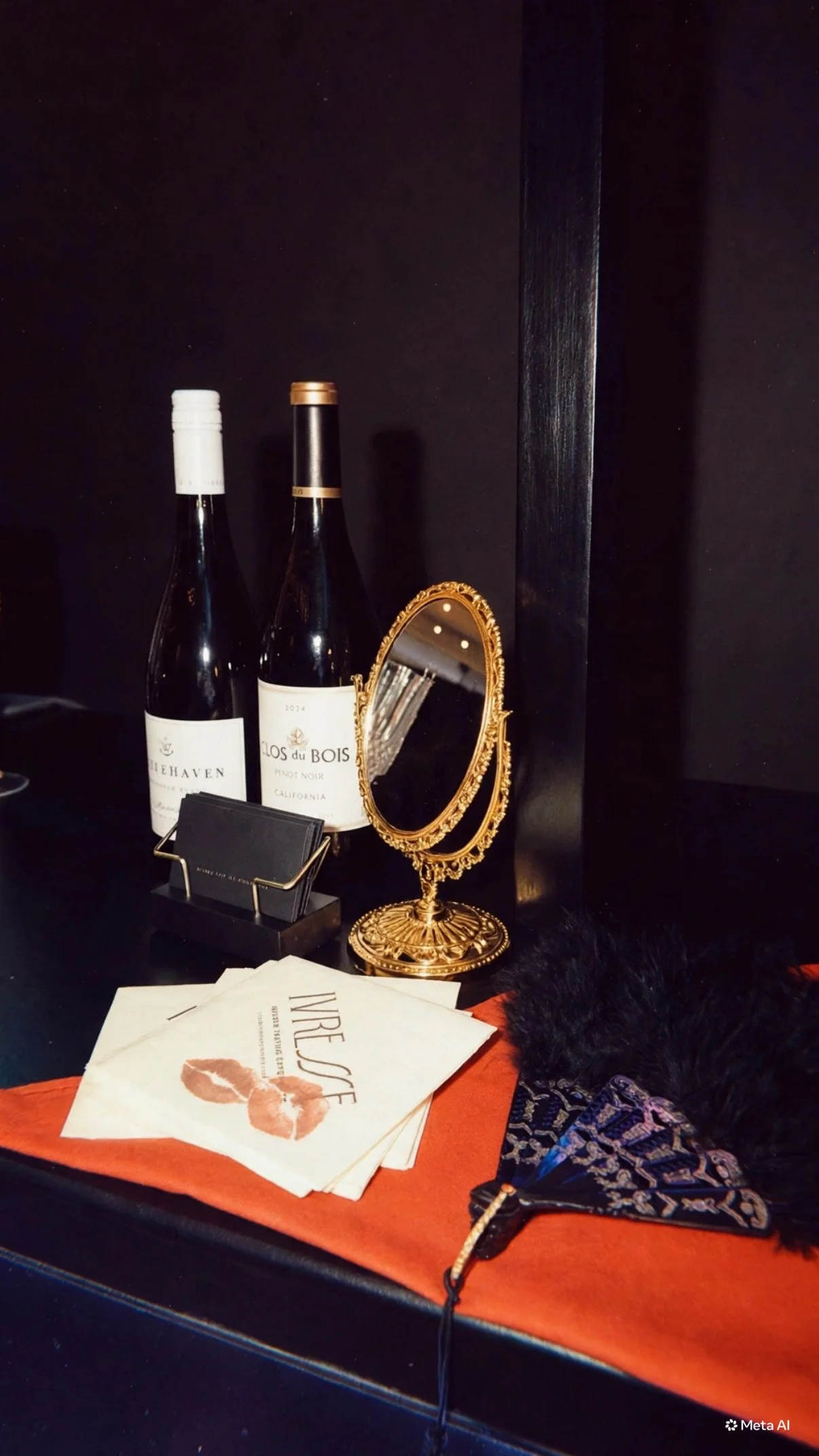 A small ornate gold mirror, two bottles of wine, and napkin with lipstick prints on red tablecloth on a black table.