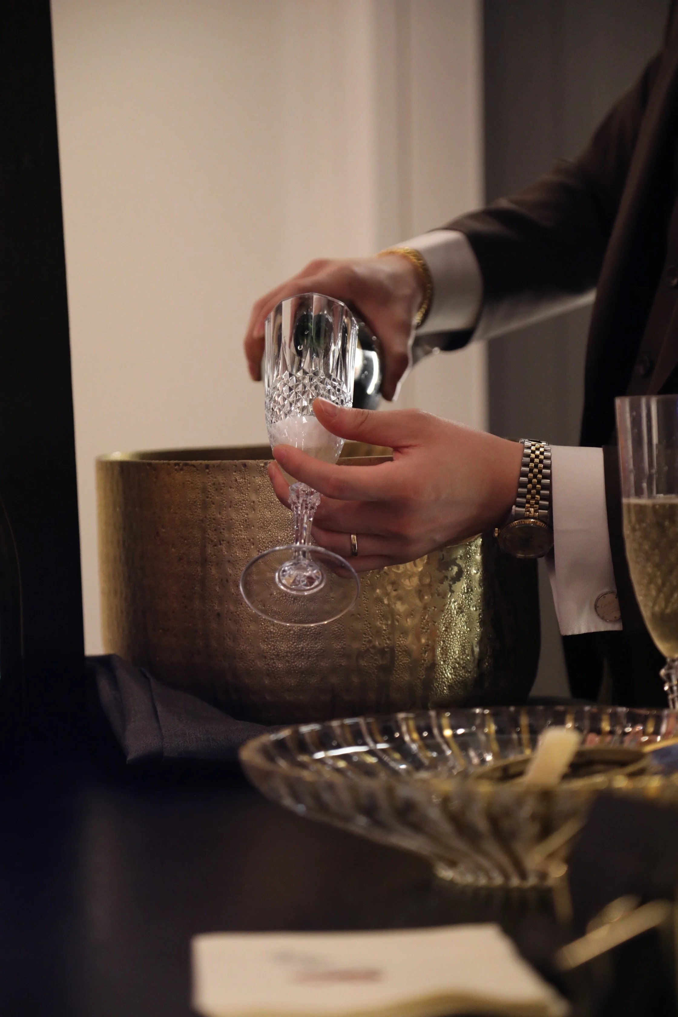 Person in formal attire pouring a drink into a crystal glass at a black and gold table setting.