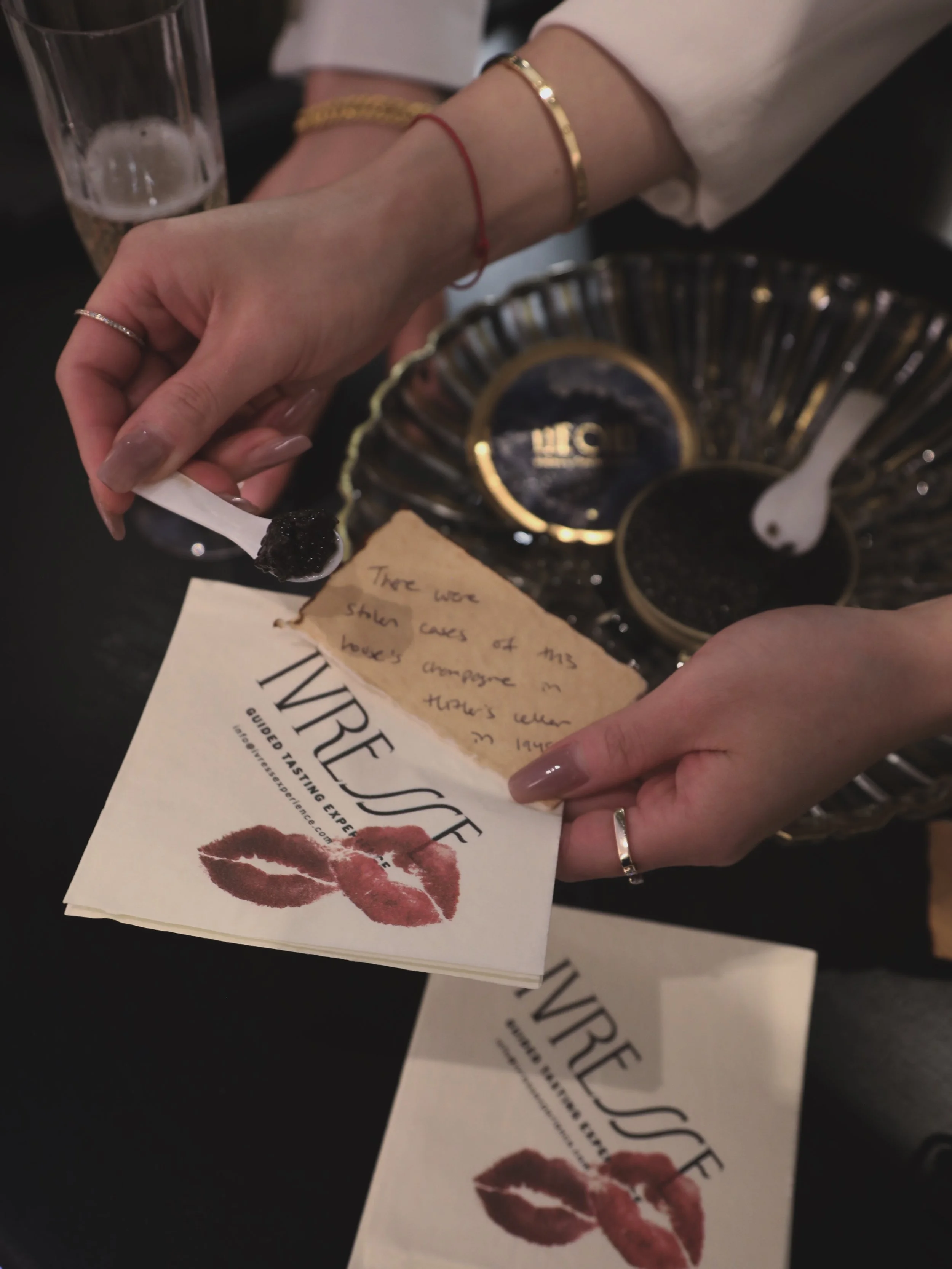 Hands holding a branded napkin with a lipstick kiss mark and a handwritten tasting note, near a decorative bowl displaying caviar, a pearl spoon with caviar, and a glass of Champagne.