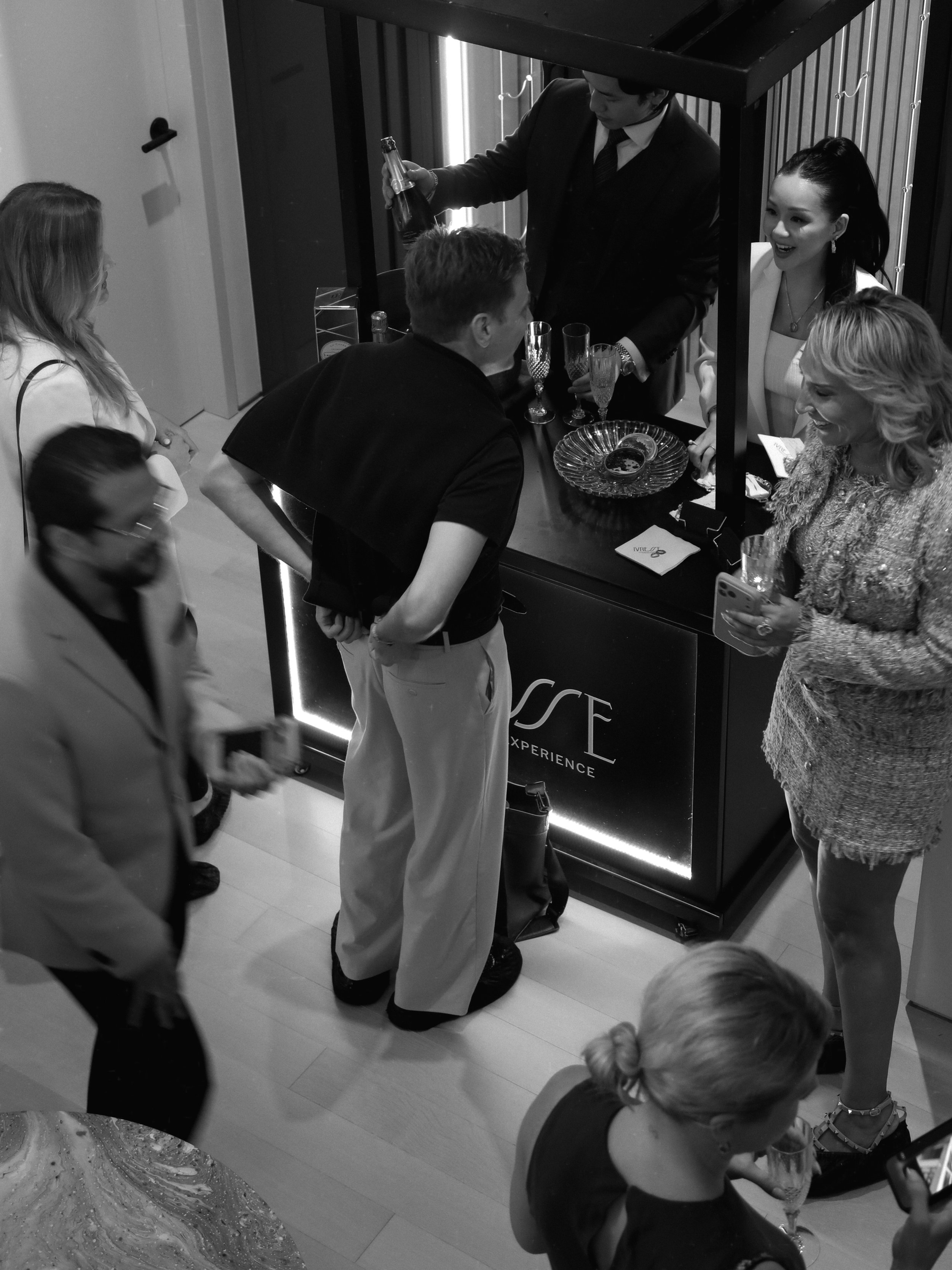 Group of people at a social gathering, some engaging in conversation, others observing, with a man serving drinks at a mobile bar or reception area. A $45M townhouse grand opening event with Nicole Gary real estate team in New York.