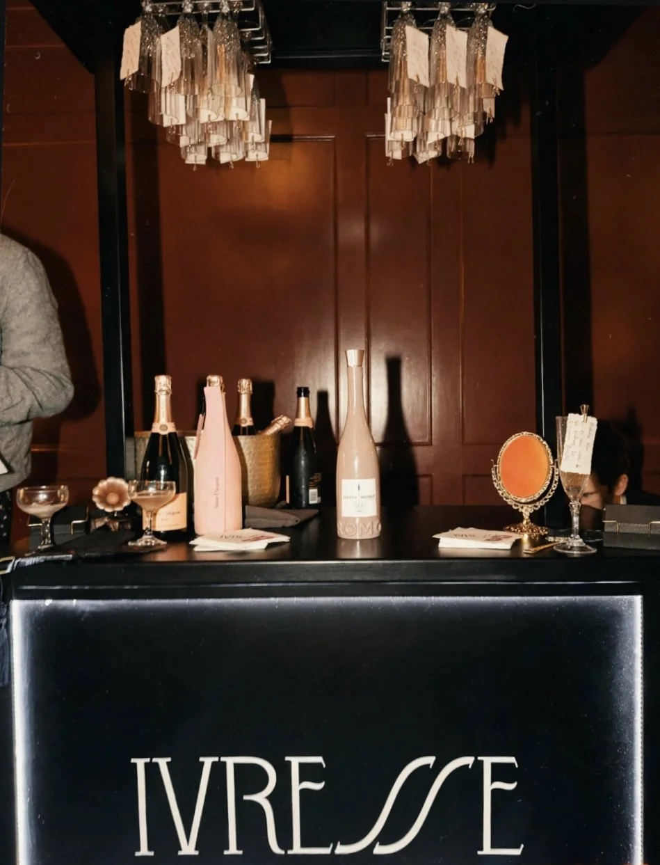 A bar with various bottles of champagne and wine, a small decorative mirror, and a black mobile bar with the word 'IVRESSE' written on it, set against a wooden wall with two chandeliers hanging overhead.