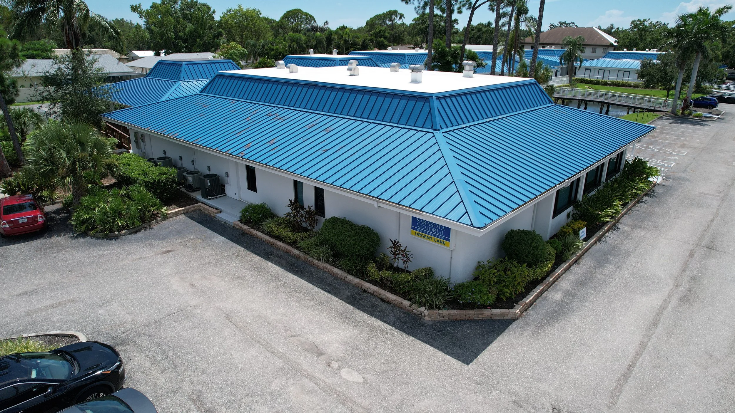 An aerial view of a single-story building with a bright blue metal roof, surrounded by parking lots, greenery, and tall trees in a sunny area. The building has a sign that reads 'Sarasota Memorial Urgent Care.'