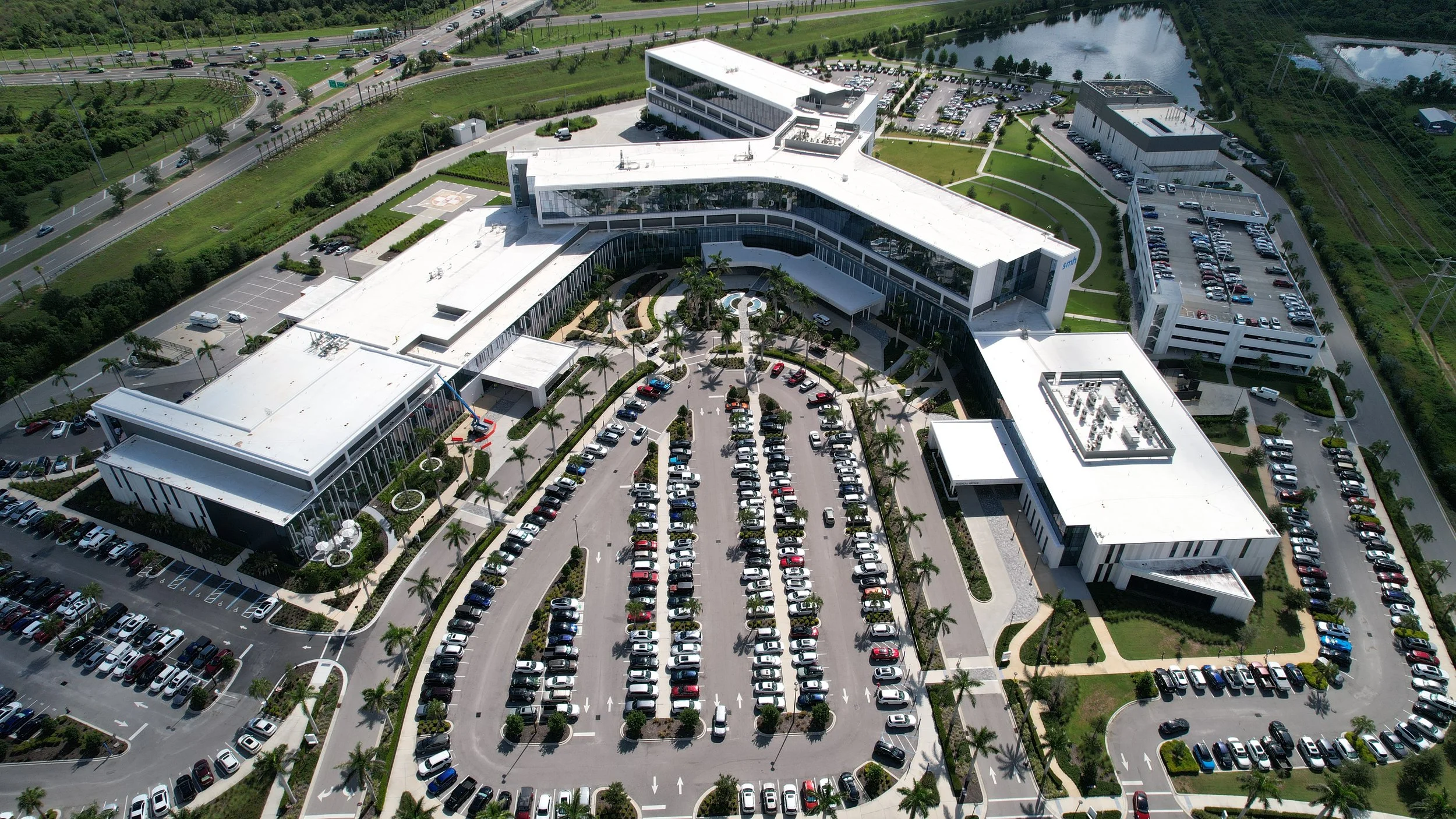 An aerial view of a large modern office complex with multiple connected buildings, parking lots filled with cars, green landscaped areas with palm trees, a water body, and surrounding roads.