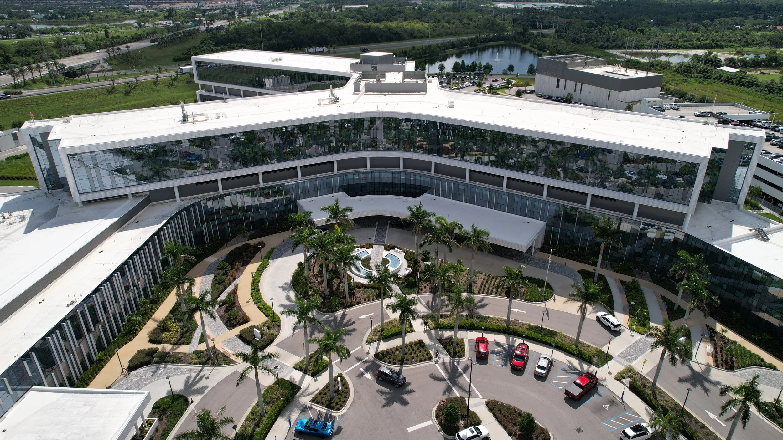 Aerial view of a modern office building with glass windows, surrounded by a landscaped area with palm trees, parking lot with cars, and a pond in the background.