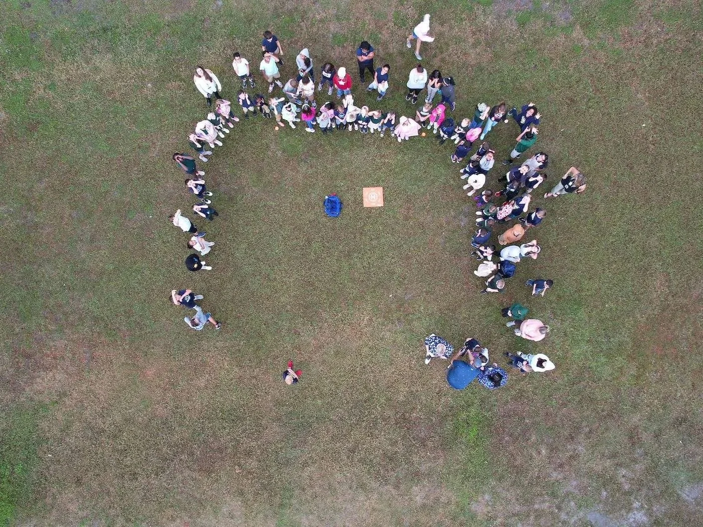 An aerial view of a large group of people forming the shape of a question mark on grass, with a small square object and a backpack near the center.