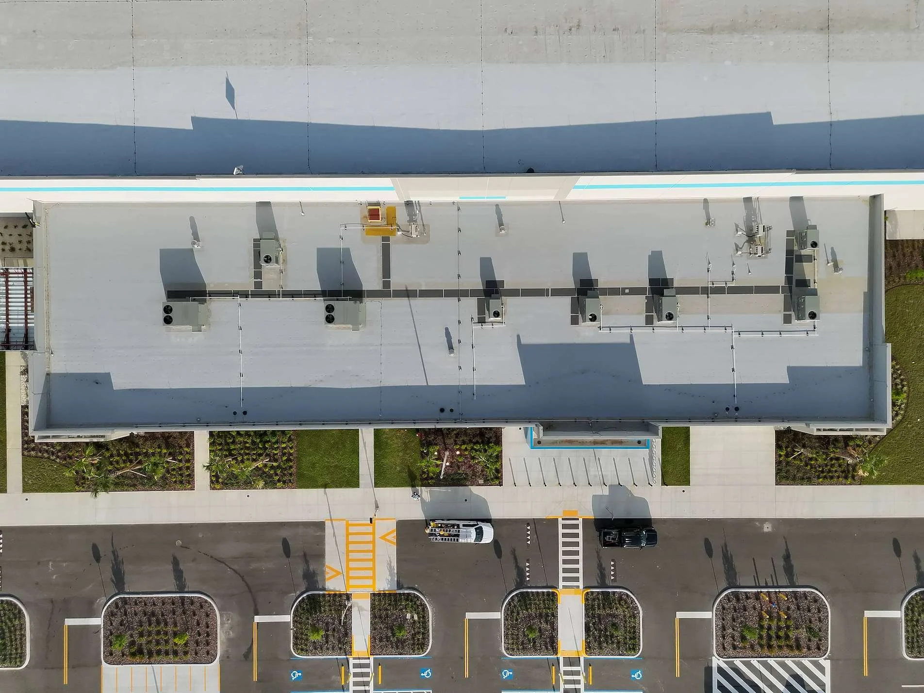 An aerial view of a building's flat roof with HVAC units and electrical equipment, with parking spaces and a street below, including handicap parking spots and a crosswalk.