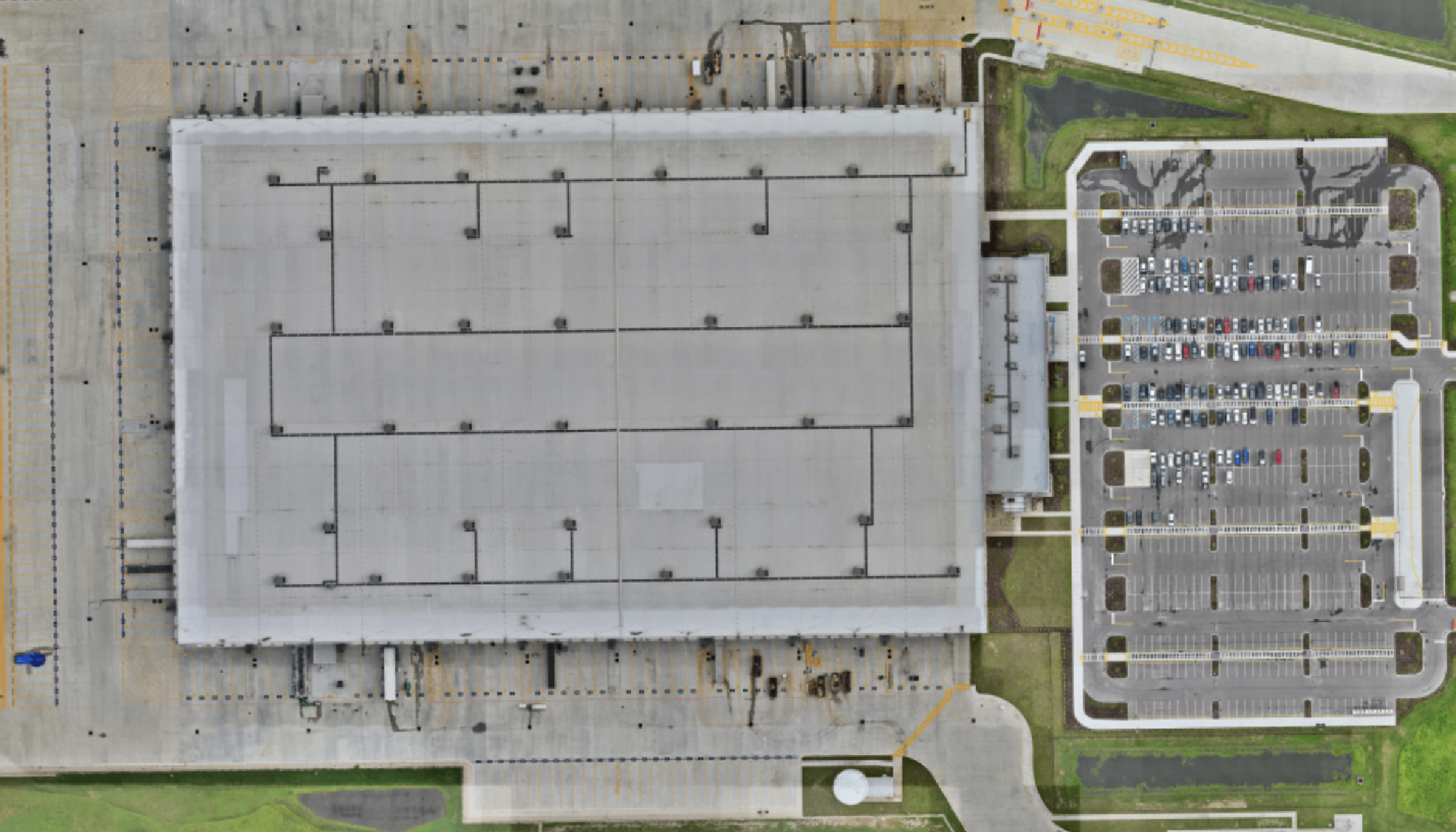 An aerial view of a large commercial building with a flat roof, surrounded by a parking lot with many marked parking spaces and cars. Green grass and small landscaped areas are visible around the parking lot.