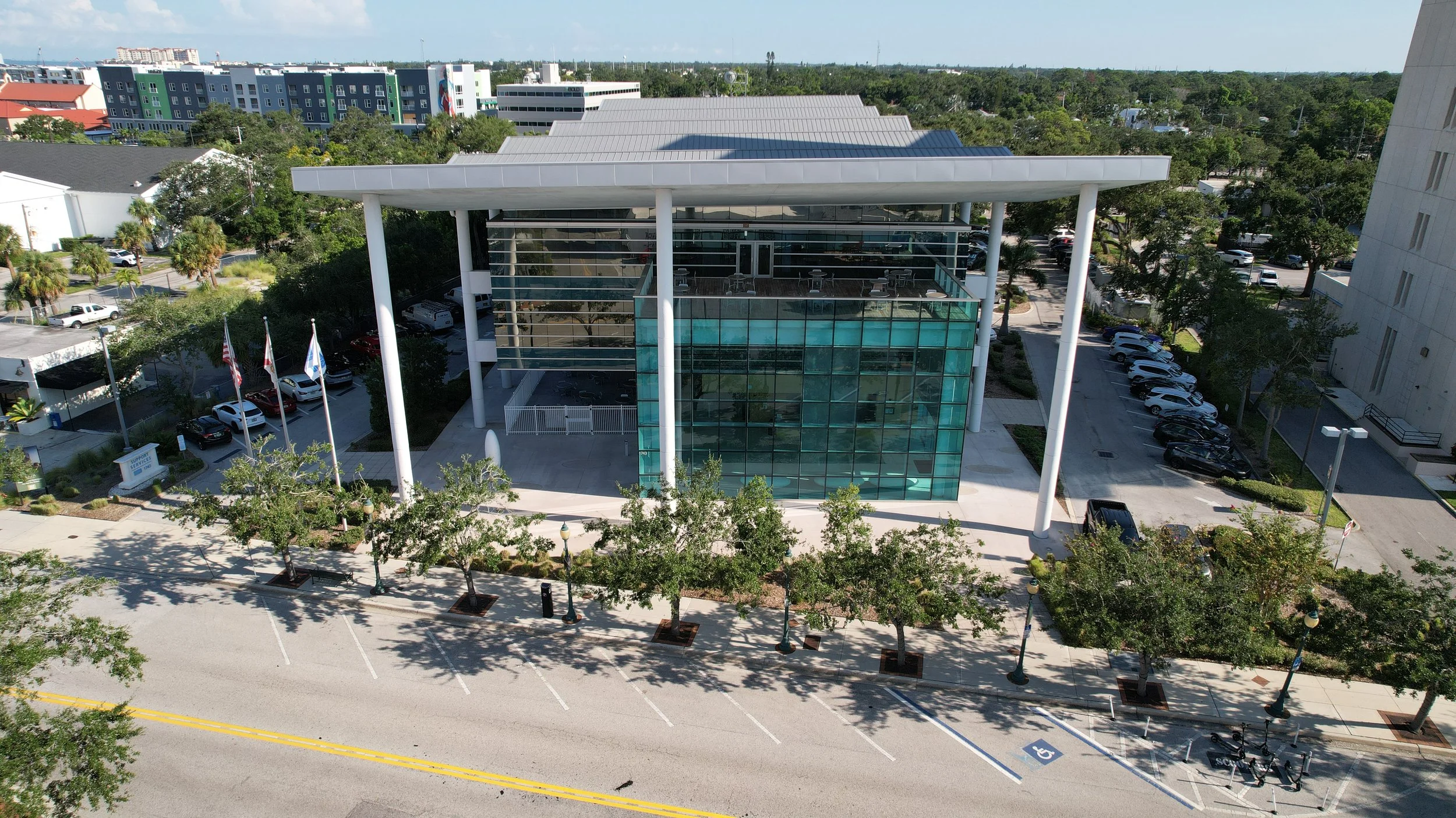 Aerial view of a modern glass-fronted building with a rooftop terrace and columns, parking lot with cars, and a surrounding cityscape including colorful apartment buildings and trees.