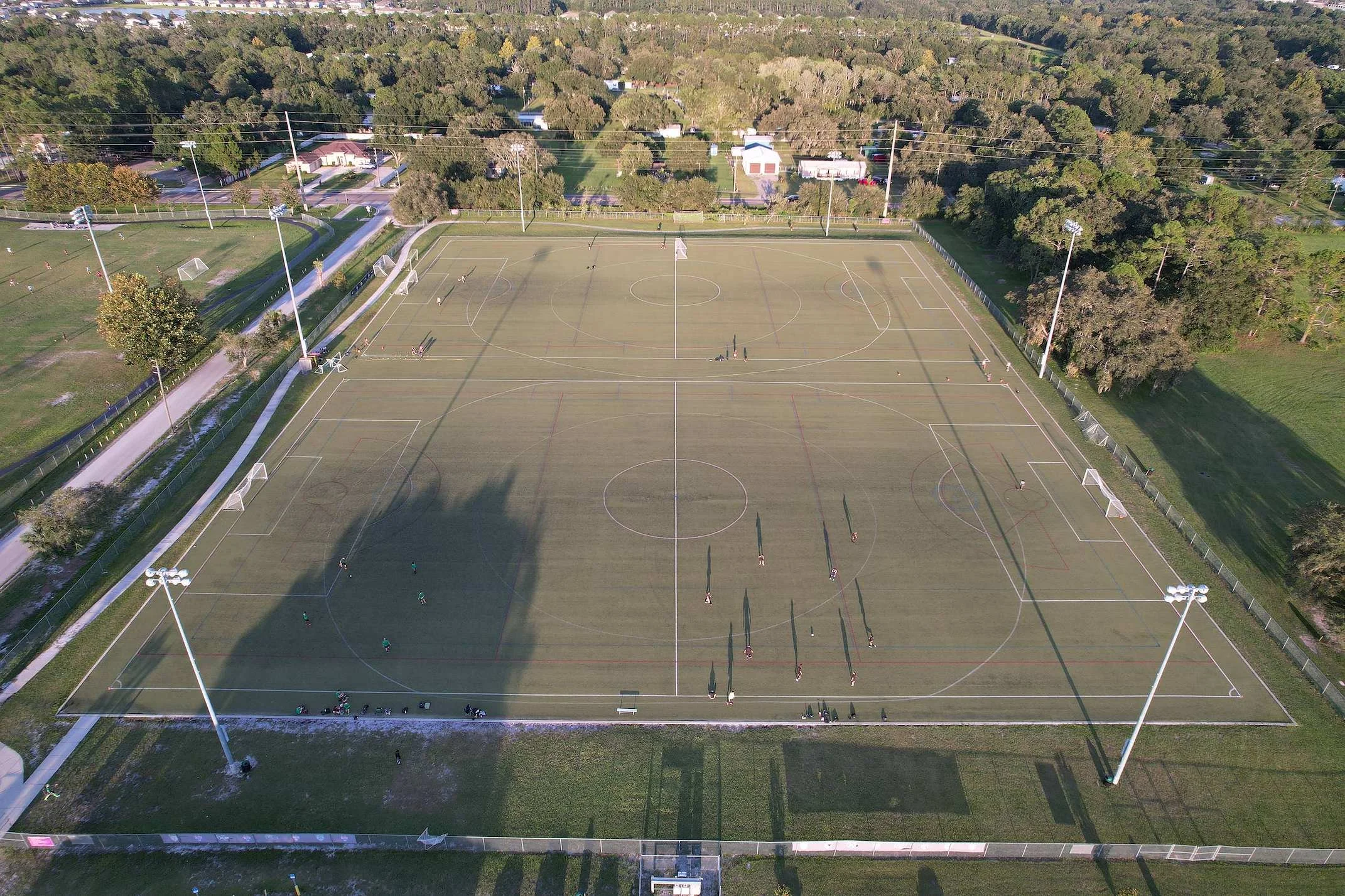 Aerial view of an empty outdoor sports field with multiple soccer goals, surrounded by tall lights, trees, and nearby roads with houses in the background.