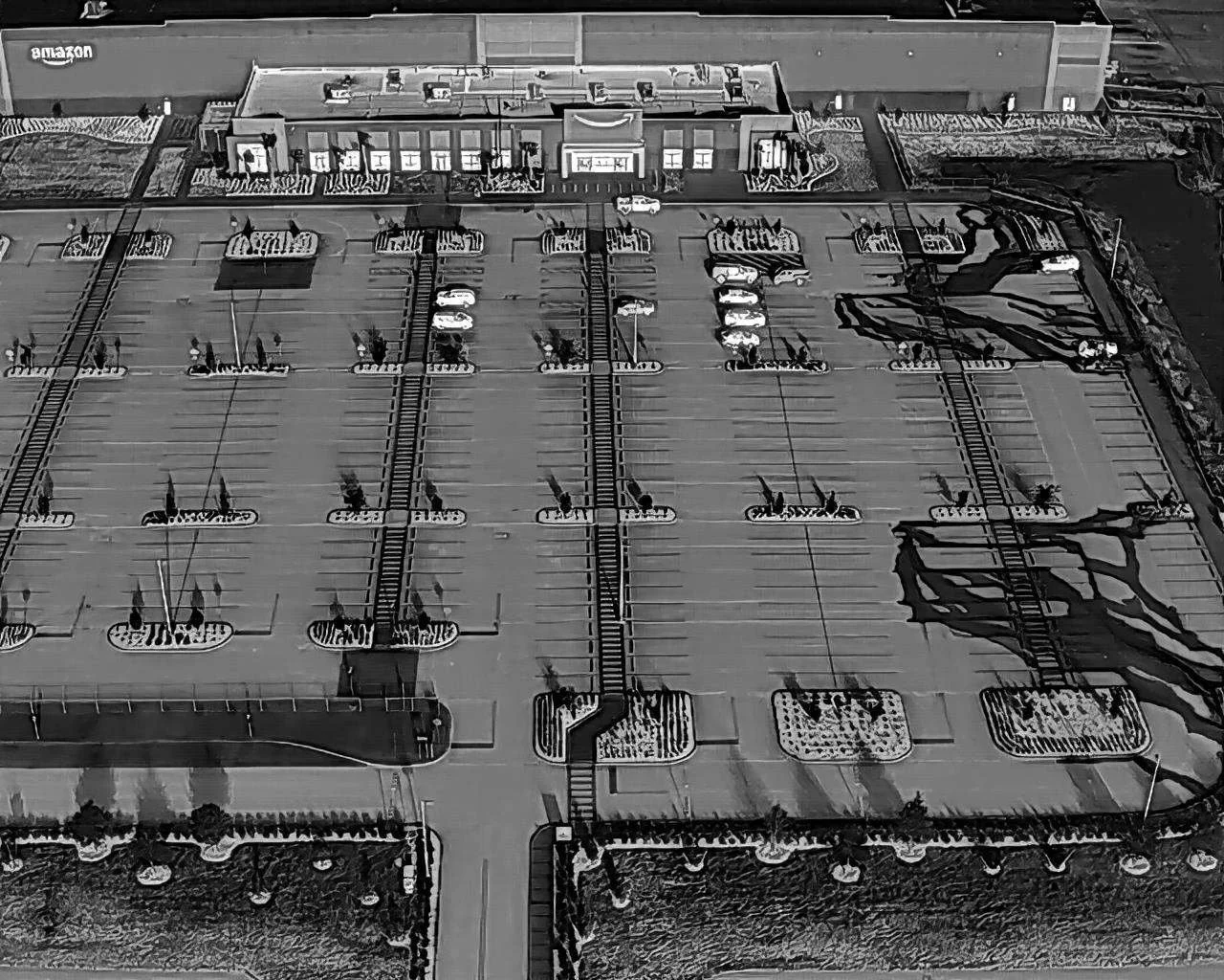 A black and white aerial view of an empty parking lot with marked parking spaces, some with small trees or plants, adjacent to a commercial building with an Amazon sign on the wall. Several parked cars are visible, and the parking lot features crosswalks, rail tracks, and landscaped areas with bushes and trees.