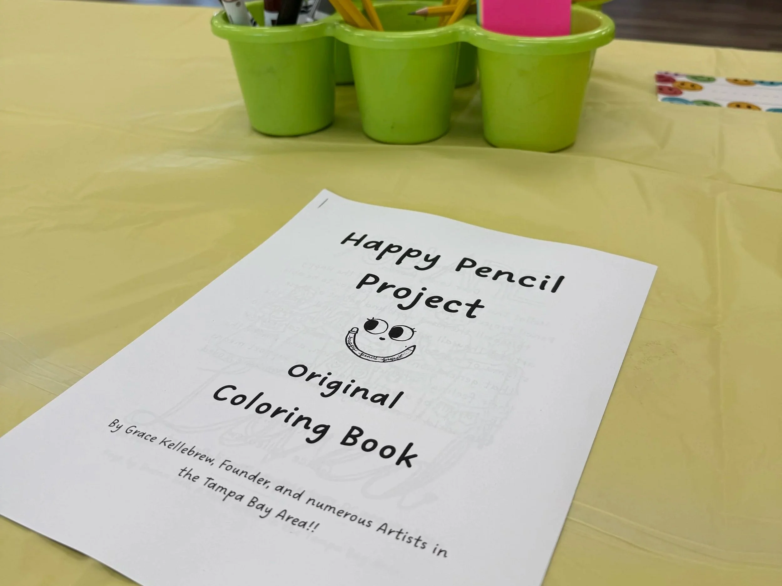 A colorful classroom activity sheet titled "Happy Pencil Project" with a smiley face drawing and bold text, lying on a yellow table next to bright green plastic containers filled with pencils and markers.