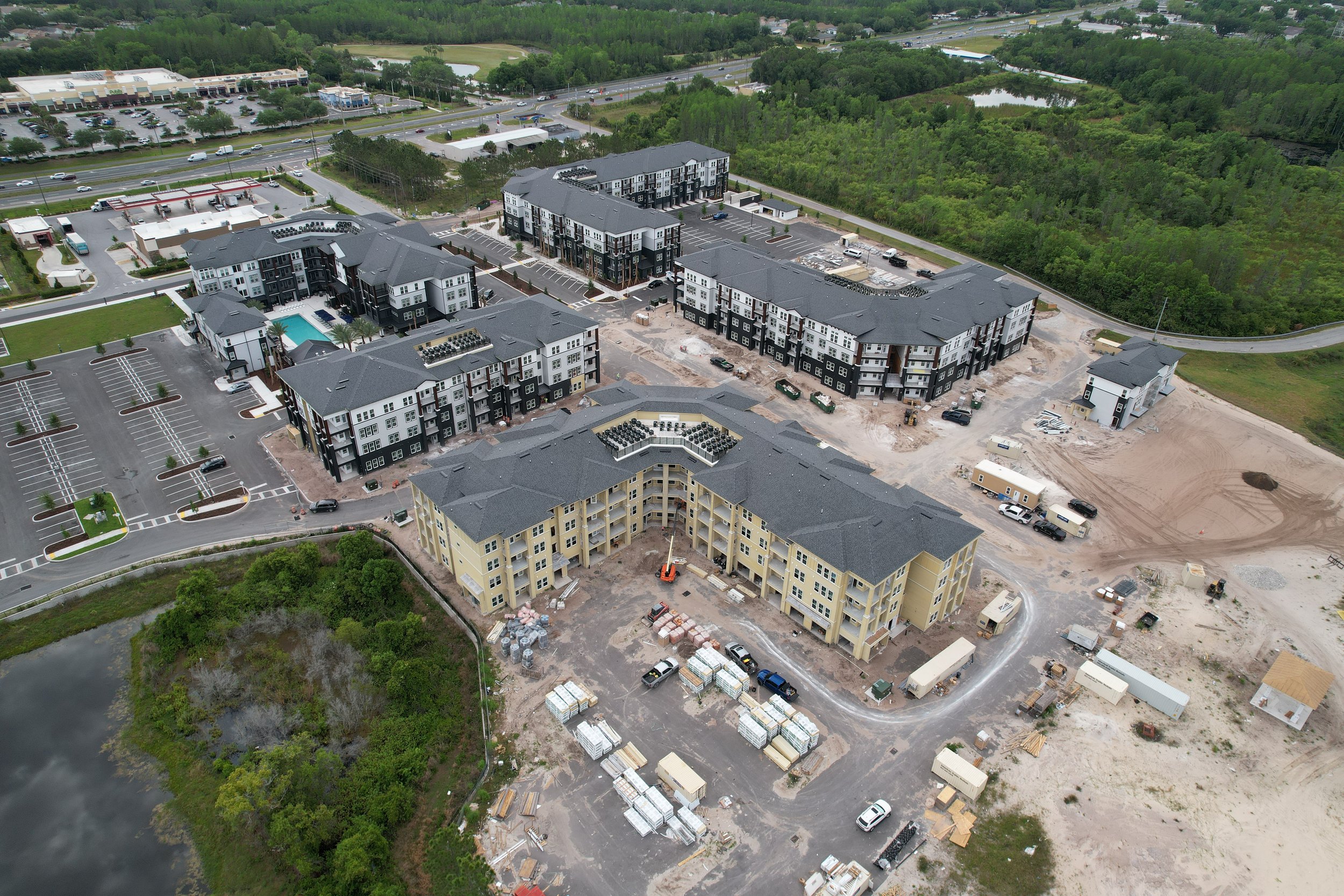 Aerial view of a large construction site with several multi-story apartment buildings in various stages of completion, surrounded by parking lots, construction materials, and some vehicles, with greenery, roads, and a small pond in the background.