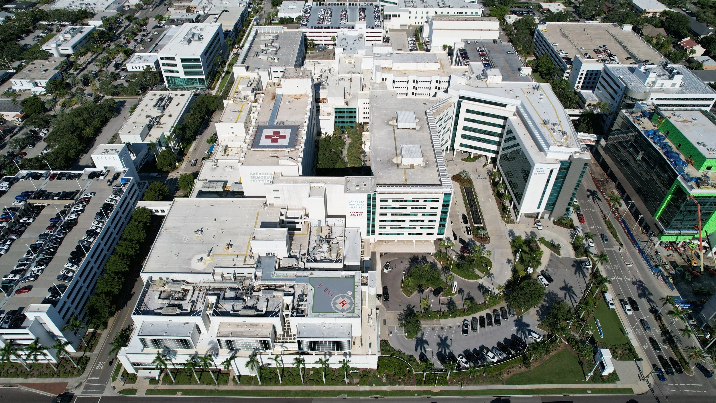 An aerial view of Sarasota Memorial Hospital with multiple buildings, parking lots, tree-lined streets, and a helipad with a red cross in the center.
