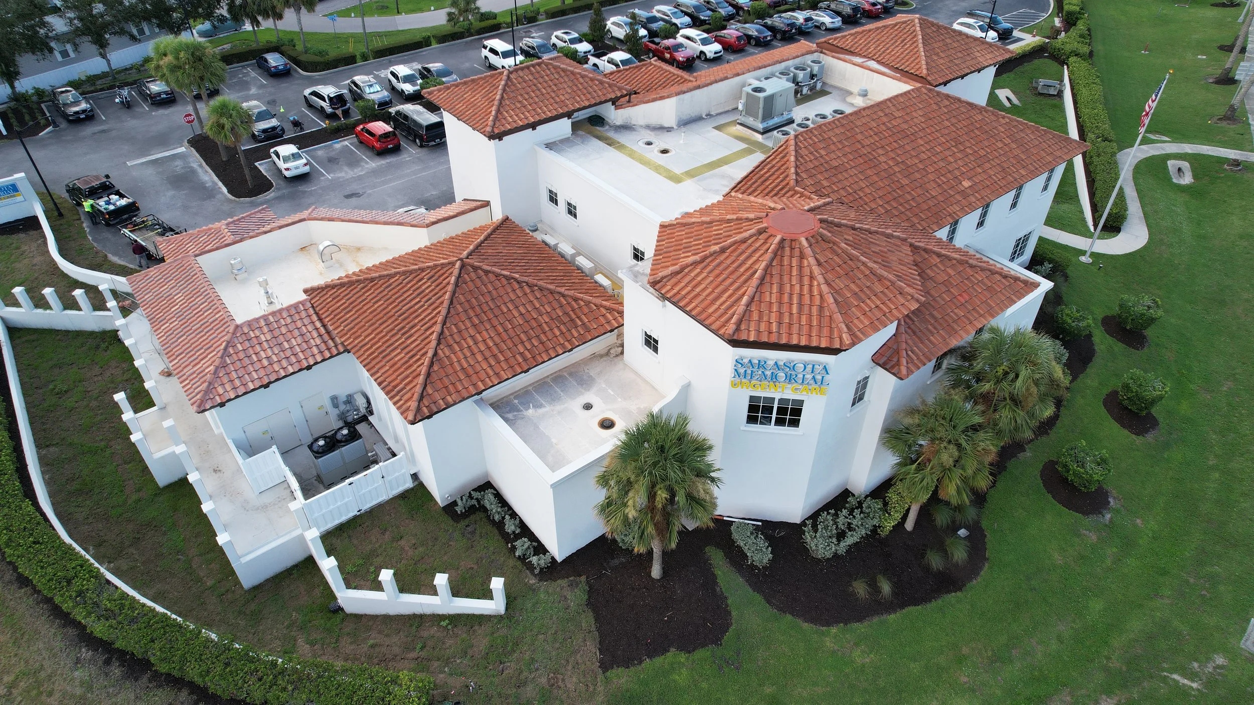 An aerial view of a white building with red tile roofs labeled "Sarasota Memorial Urgent Care," surrounded by green landscaping, palm trees, a parking lot with cars, and a sidewalk with a small American flag.