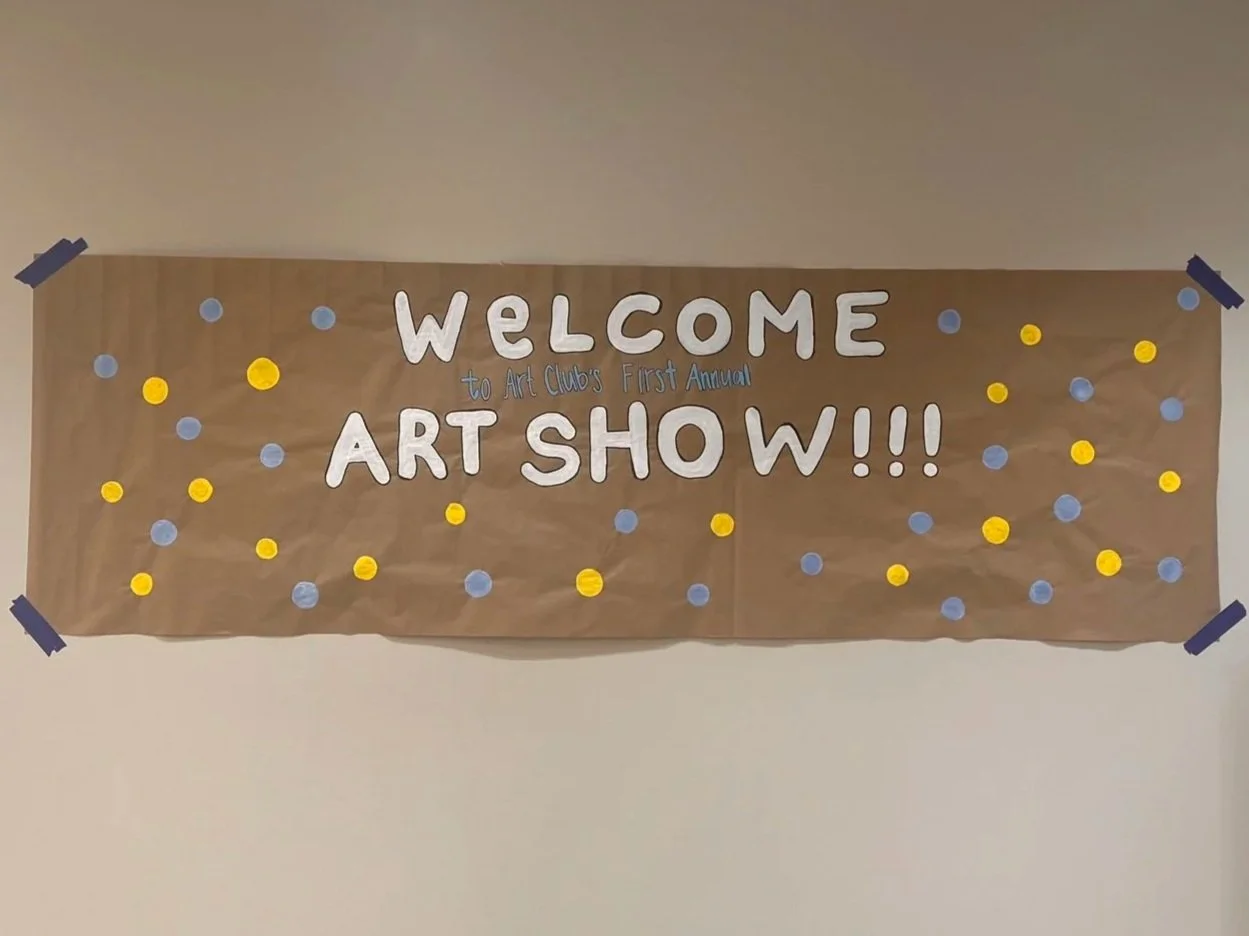 Decorative banner with the text 'Welcome to Art Club's First Annual Art Show!!!' on a brown paper background, decorated with blue and yellow paper dots, taped at the corners.