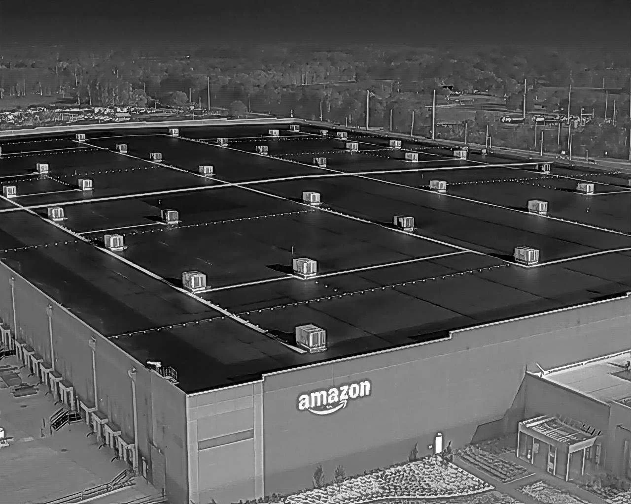 Aerial view of an Amazon distribution center with a flat roof, HVAC units, and a parking lot, in a spacious area with fields and a highway in the background.