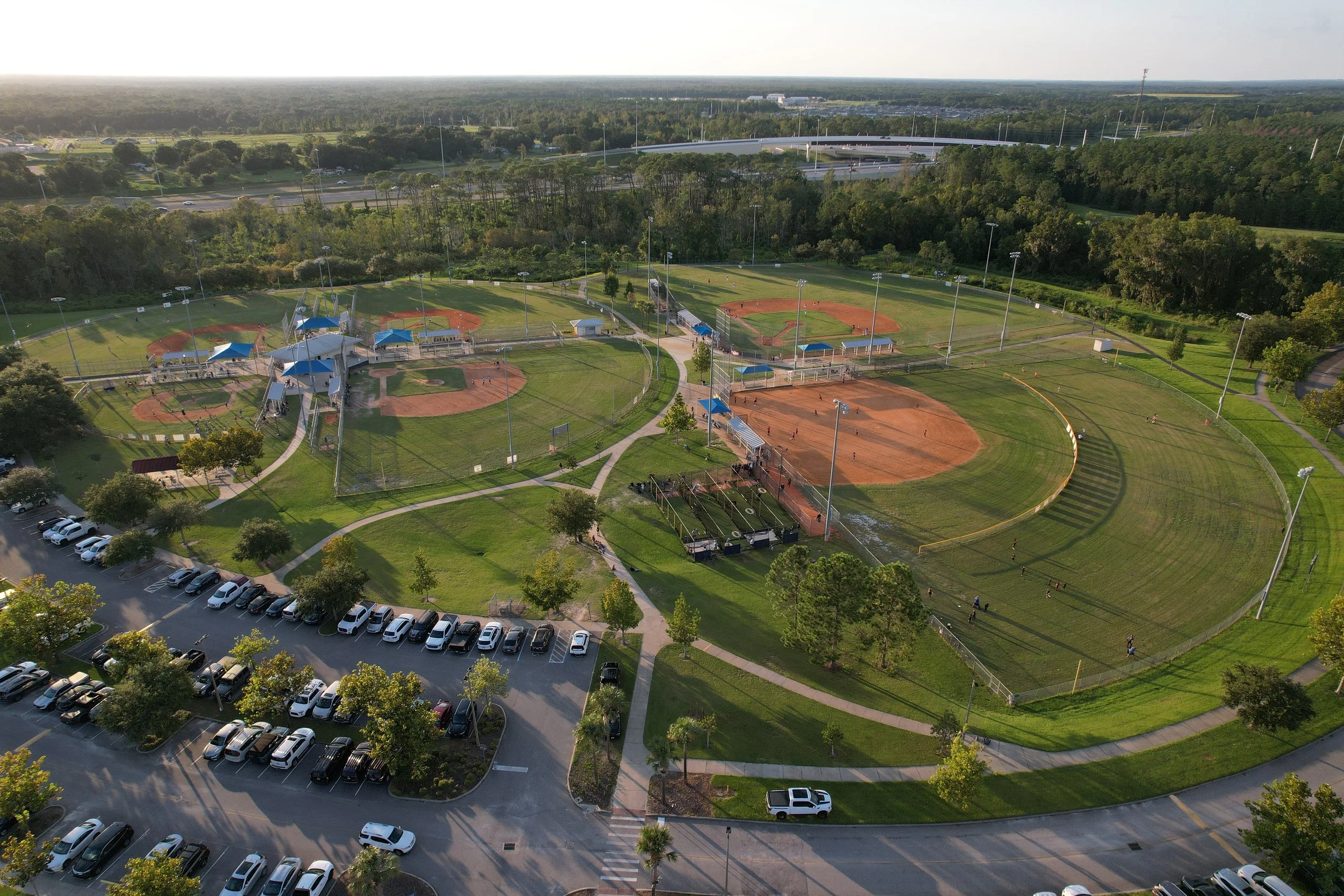 Aerial view of a baseball park with multiple fields, parking lot, and surrounding trees, with highways and urban area in the background.