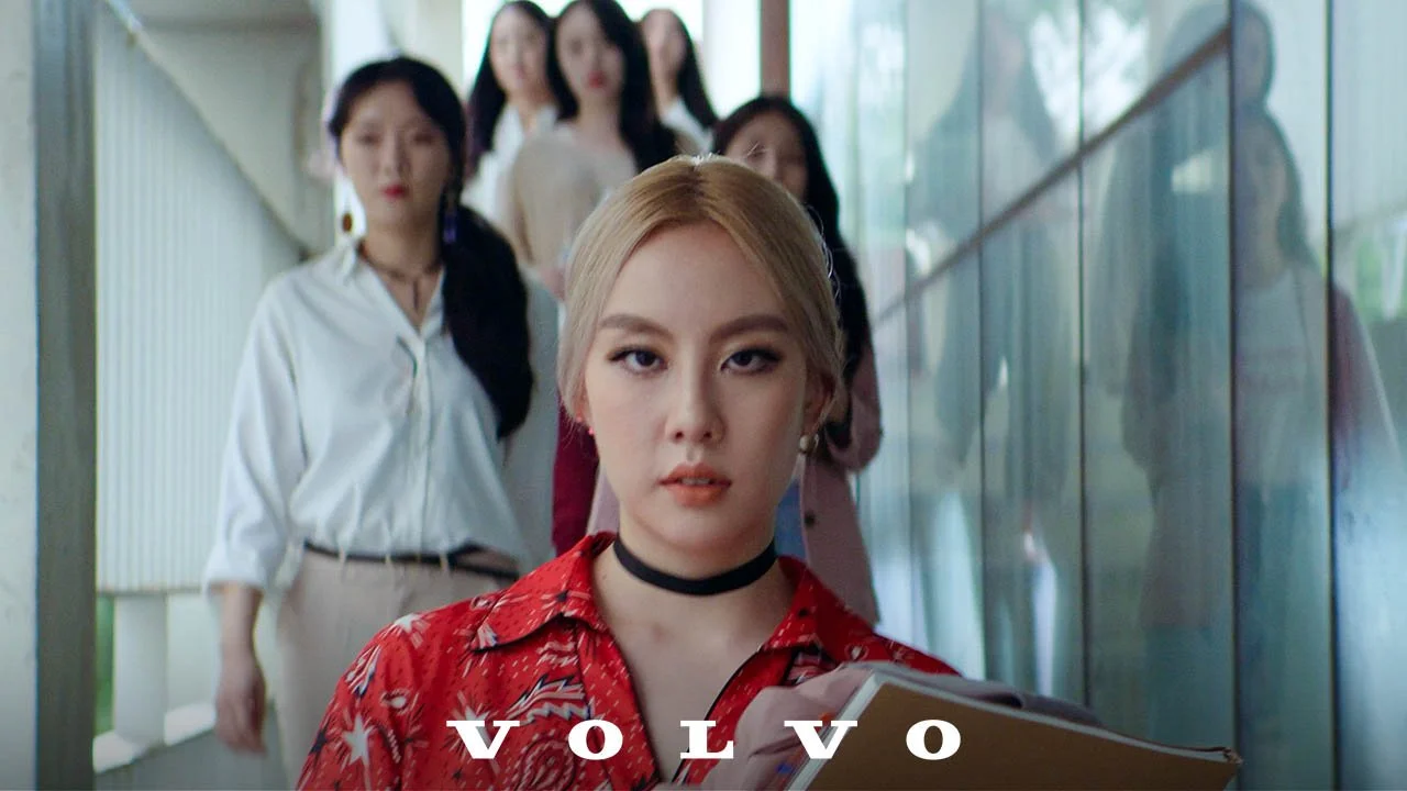 A young woman with blonde hair, wearing a red patterned shirt, choker, and earrings, leading a group of women walking behind her. The women are dressed in casual to professional attire, walking on a set of stairs beside a glass wall. The Volvo logo is visible at the bottom of the image.