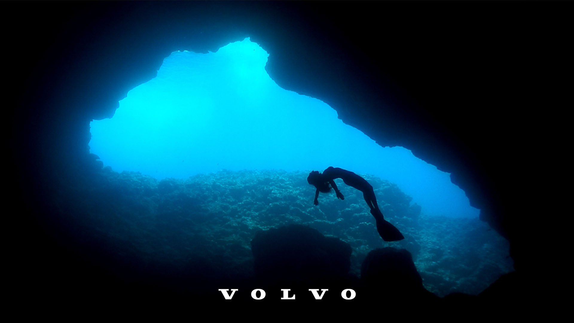 A scuba diver swimming inside a cave or underwater cavern with a bright opening above, revealing blue water and rocky formations. The word 'V O L V O' is displayed at the bottom.