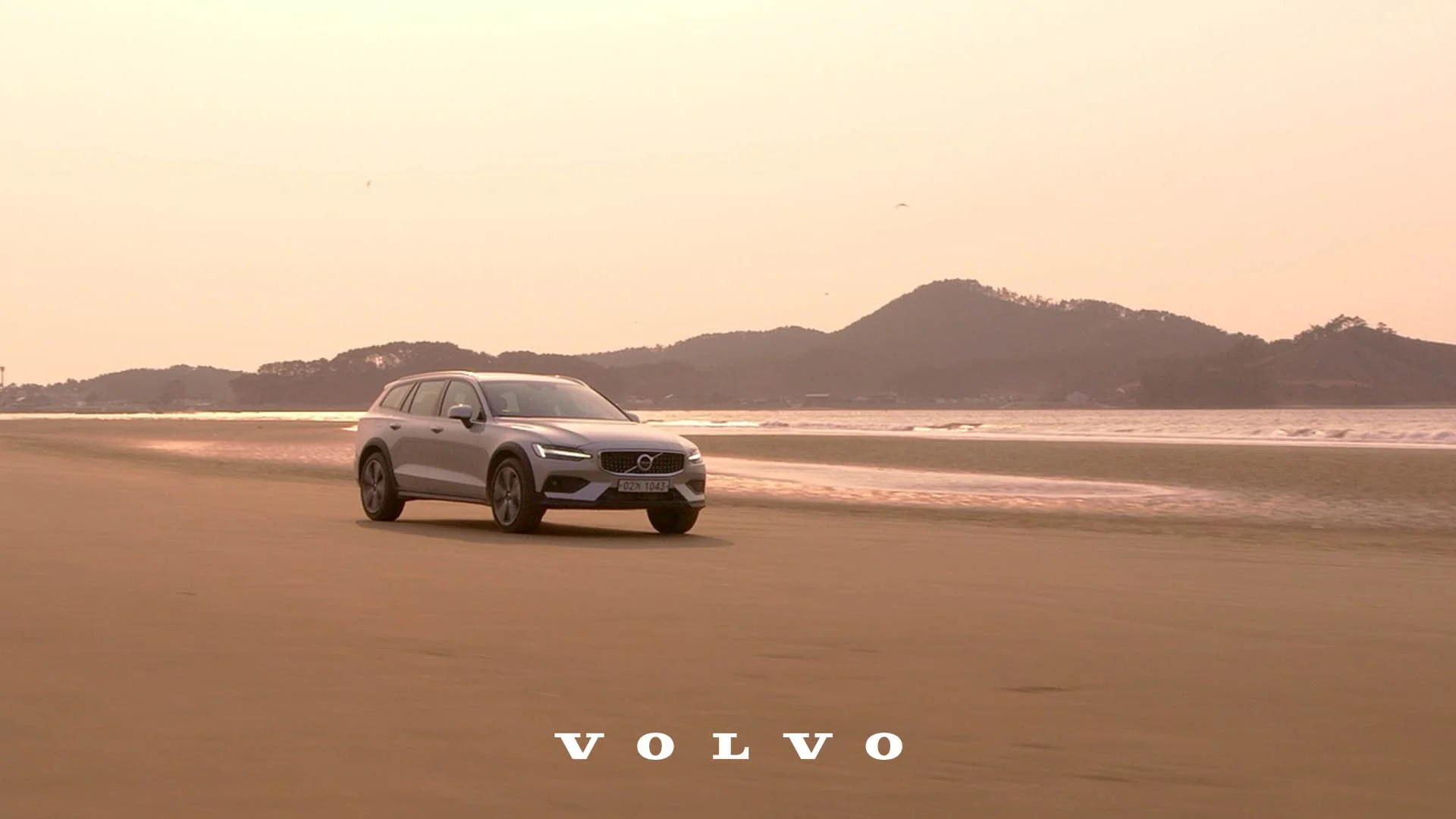A silver Volvo SUV driving on a beach road near the ocean with hills in the background during sunset.