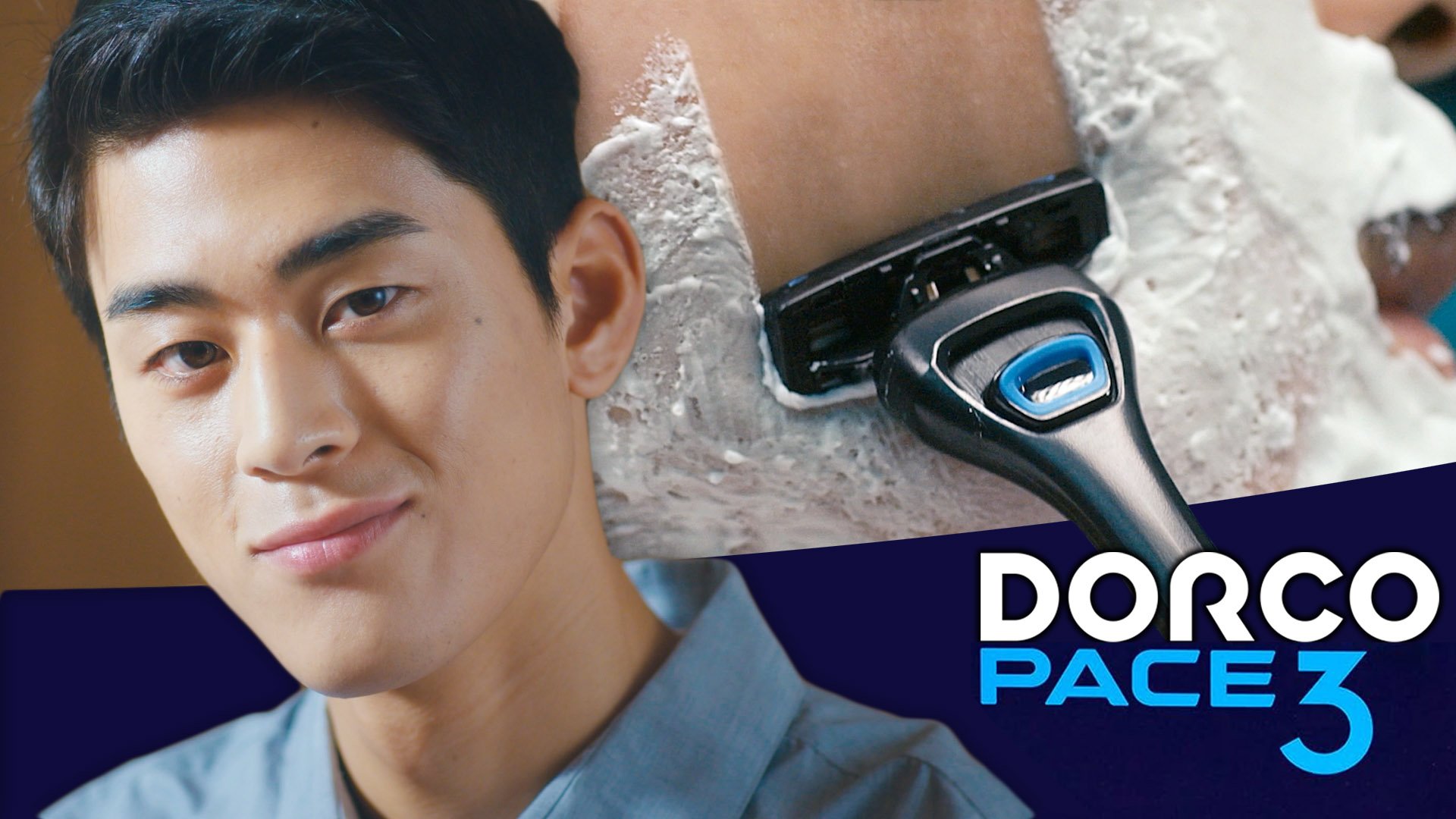 A young man with short black hair and a slight smile is getting a haircut with an electric clipper. The hair and part of the neck are shaved, with some white foam or hair product on the scalp. The background is blurred, and there is a blue section with white and blue text that says 'DORCO PACE 7'.